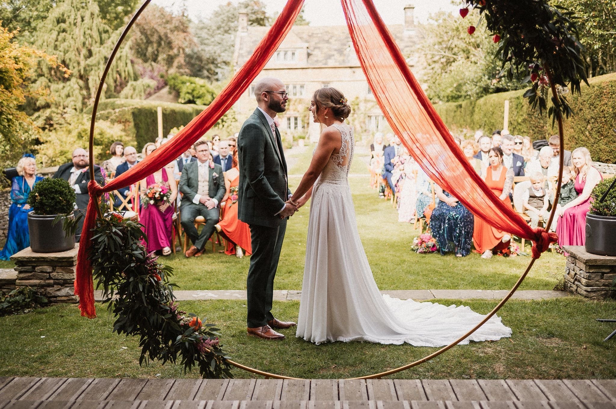 Wedding Ceremony photo showing the bride and groom in the foreground frames in a hoop draped in red cloth, and their guests sat for the ceremony in the background.