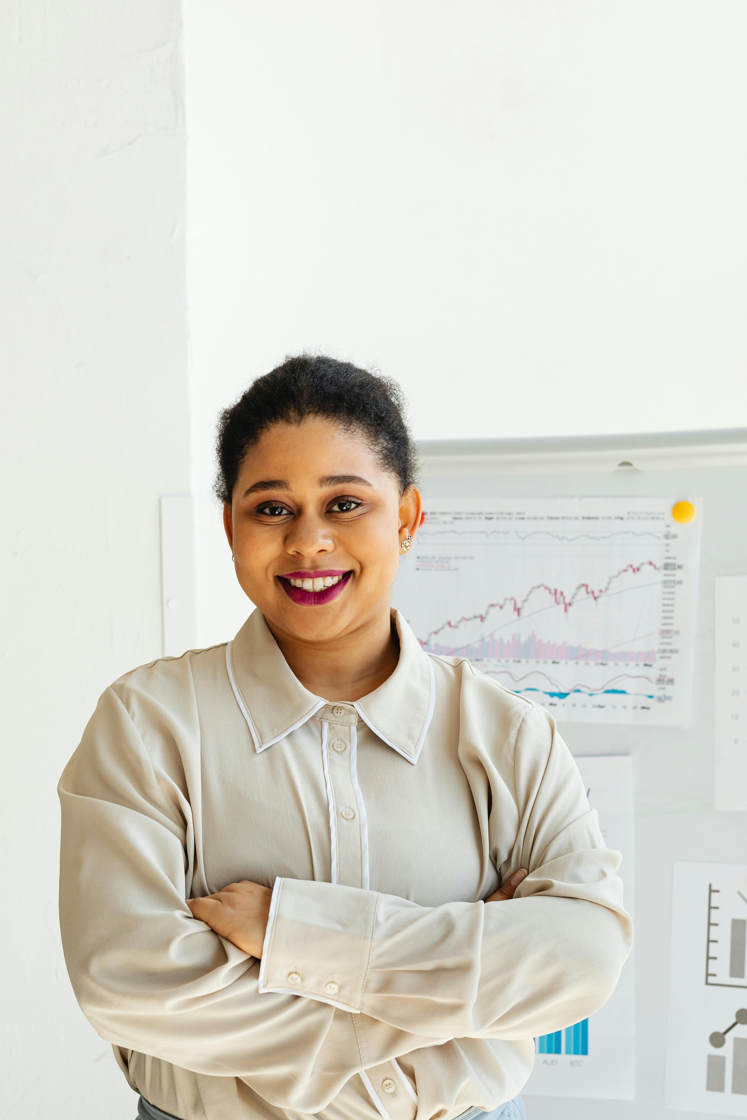 Smiling woman with dark hair tied back, wearing a beige button-up shirt, standing with arms crossed in front of a white wall with charts and graphs.