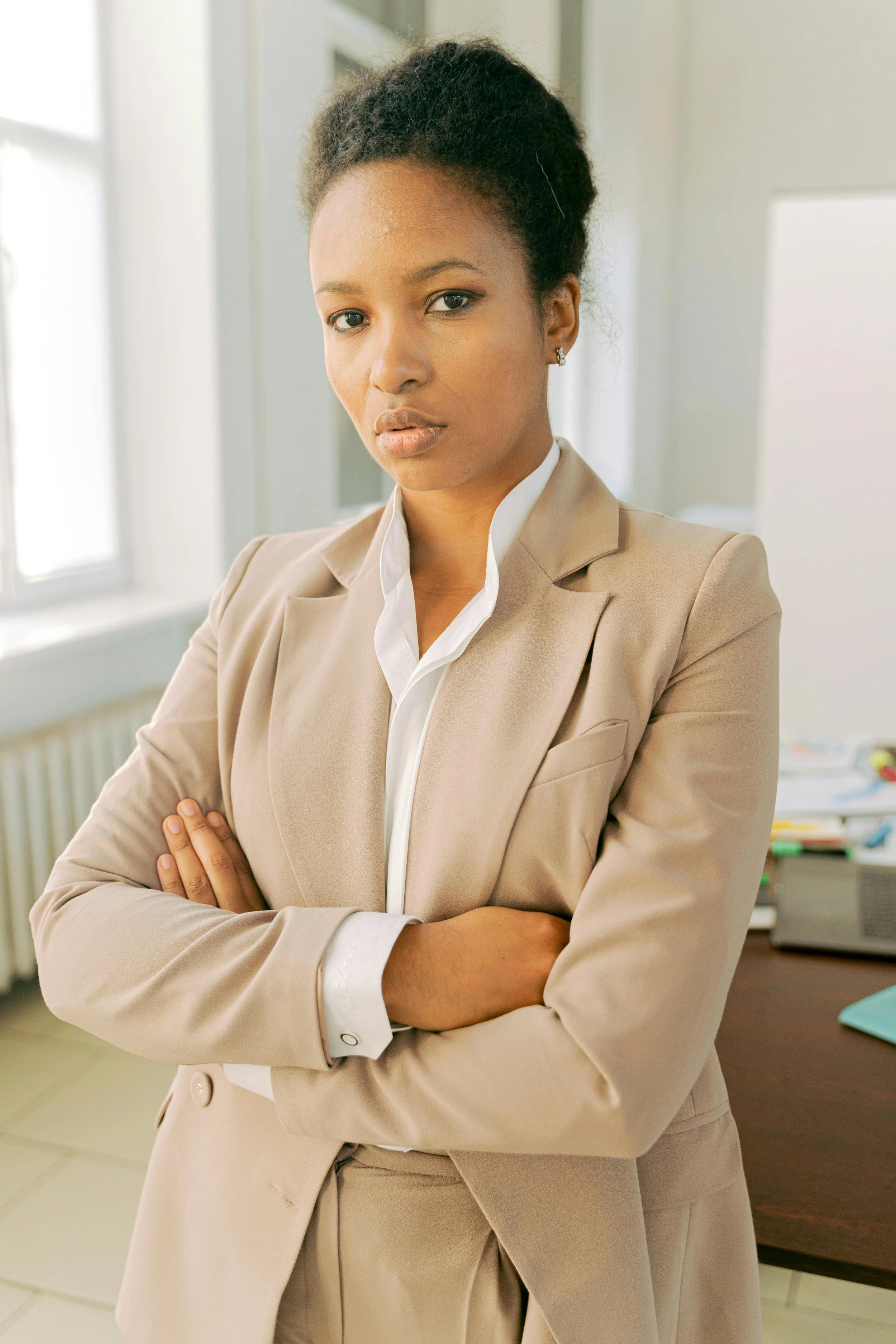 A confident woman in a beige suit crossed arms standing in an office.