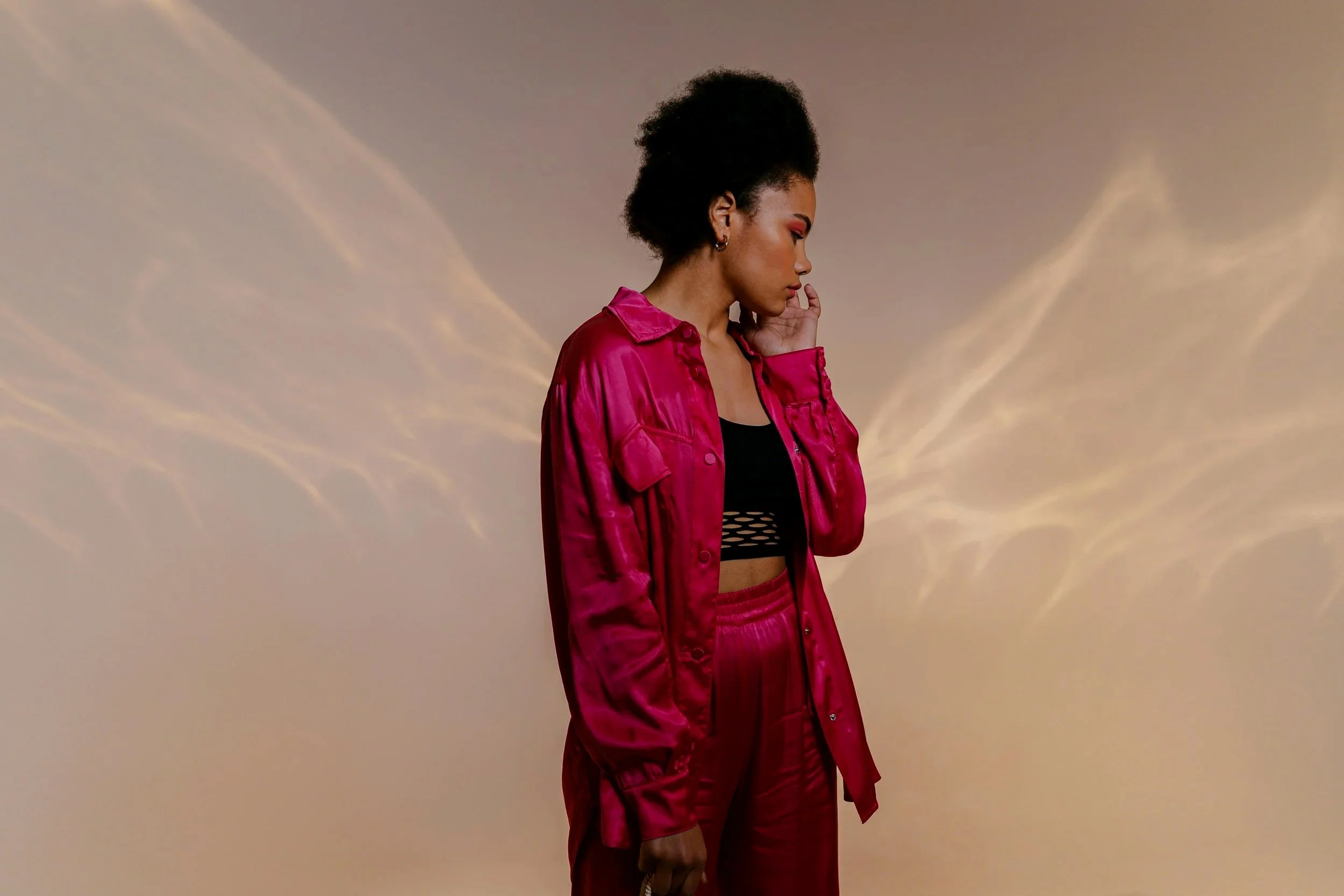A young woman with short curly hair and dark skin stands with her hand near her face, wearing a bright pink satin jacket over a black mesh top, against a backdrop of a sunset sky with wispy clouds.