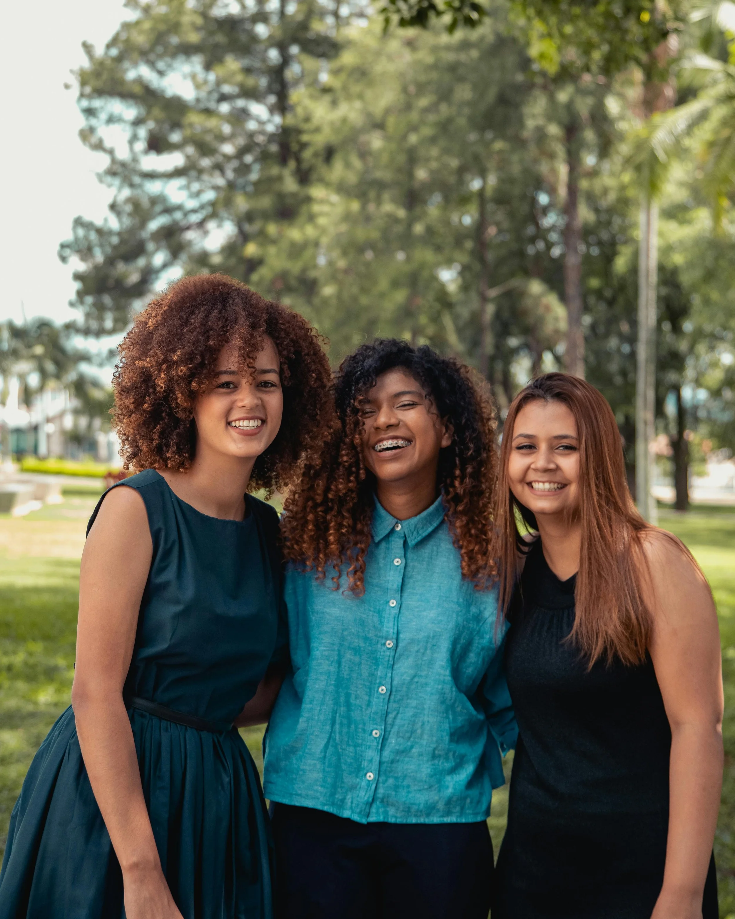 Three women standing close together outdoors, smiling, with trees in the background.