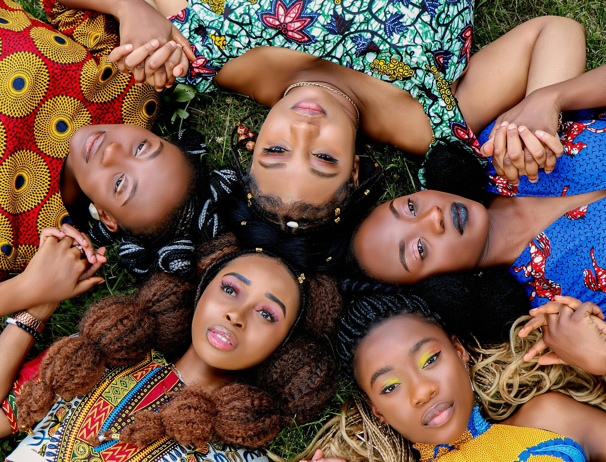 Multiple diverse women lying on the grass holding hands, dressed in colorful traditional African clothing and accessories.