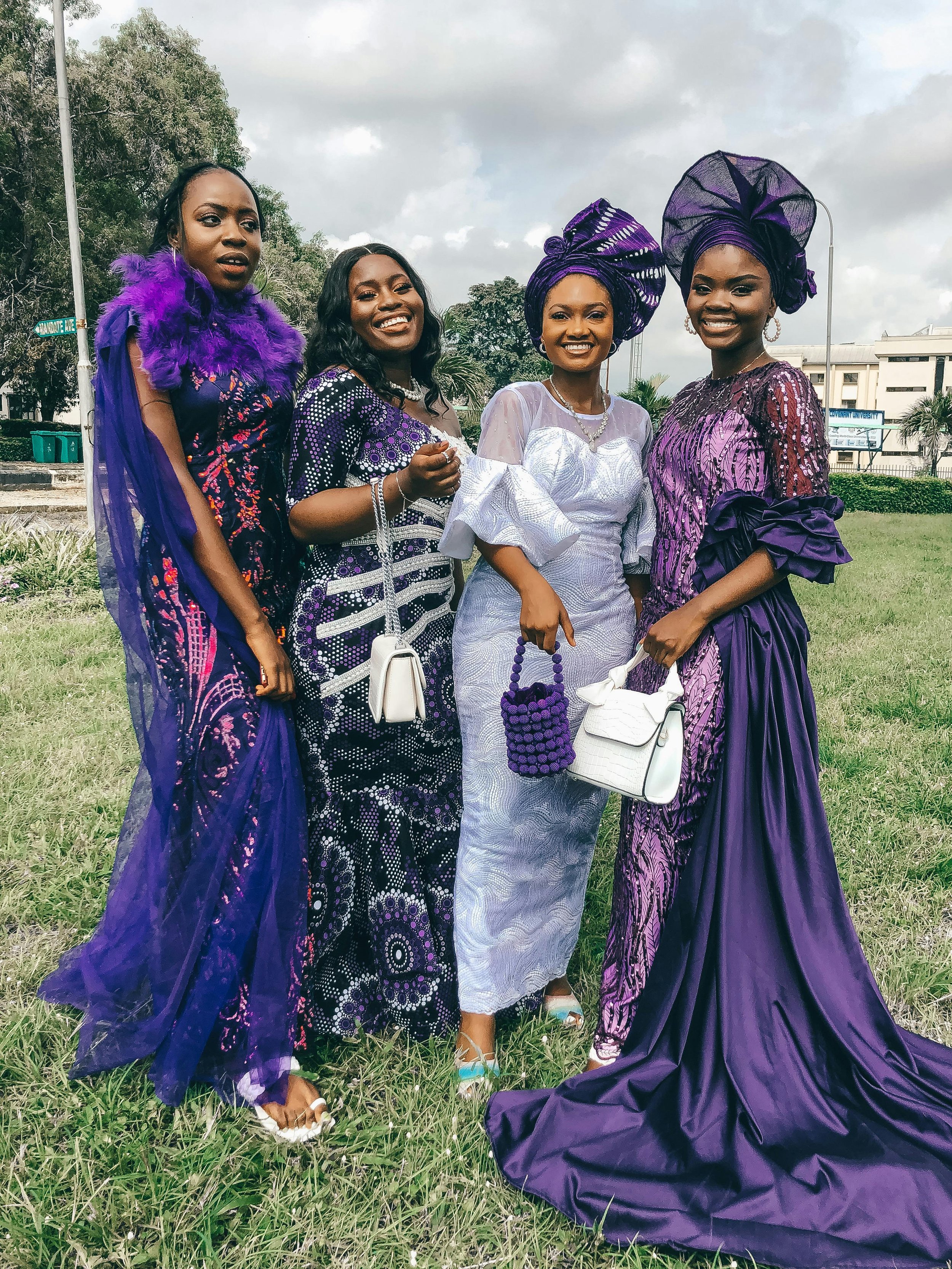 Four women dressed in traditional Nigerian attire, standing on a grassy field with trees and buildings in the background. They are wearing purple and white outfits with headwraps, smiles, and holding handbags.