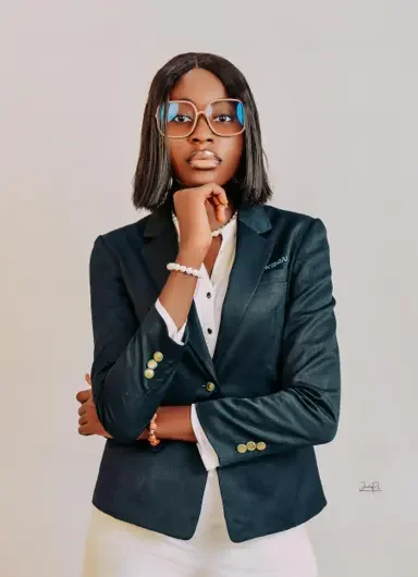 A woman with shoulder-length black hair, glasses, and dark lipstick wearing a navy blazer over a white shirt, standing against a gray background, with her hand resting on her chin.