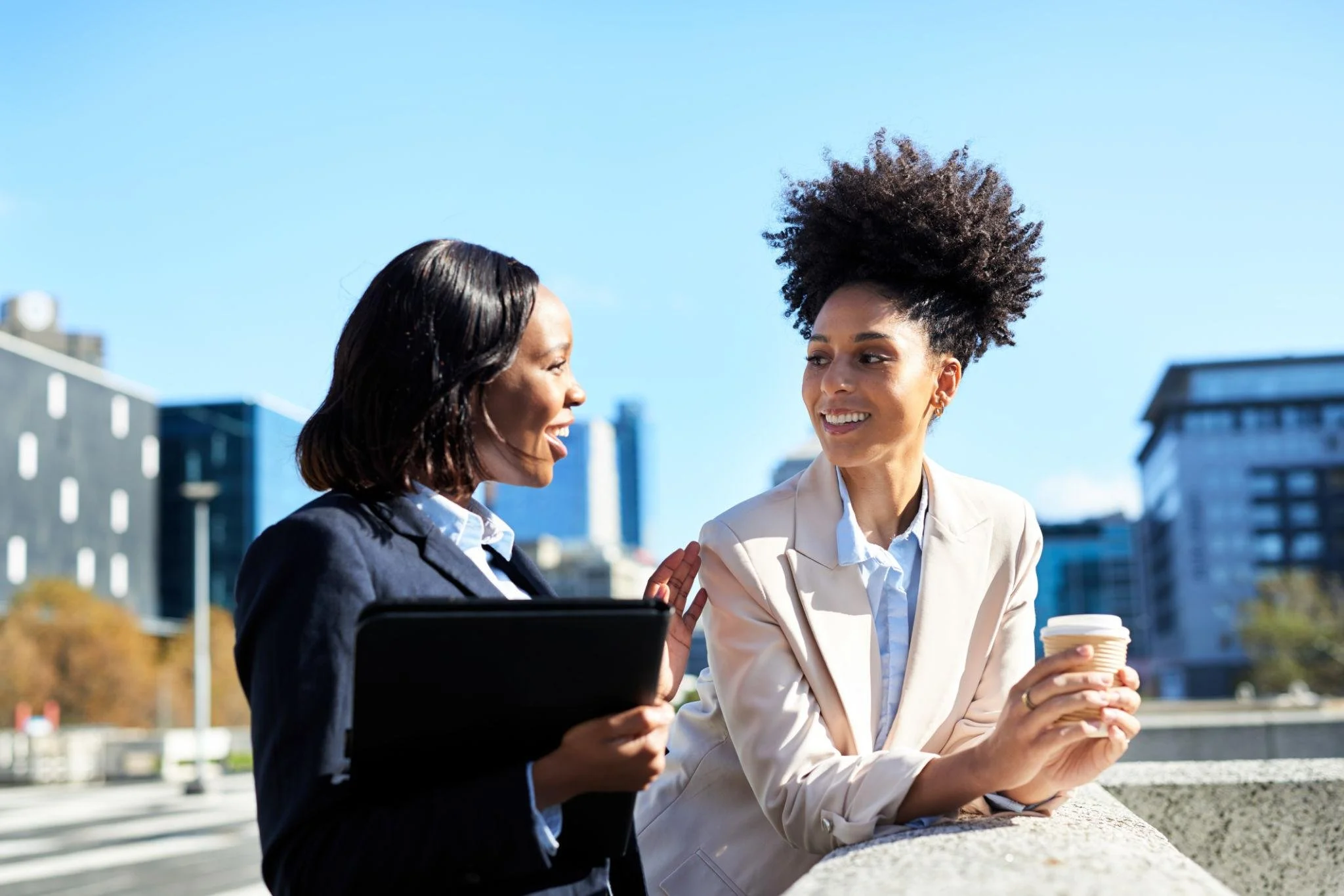 Two women are having a conversation outdoors in an urban setting, with buildings in the background. One woman is holding a disposable coffee cup while the other is holding a folder or tablet.