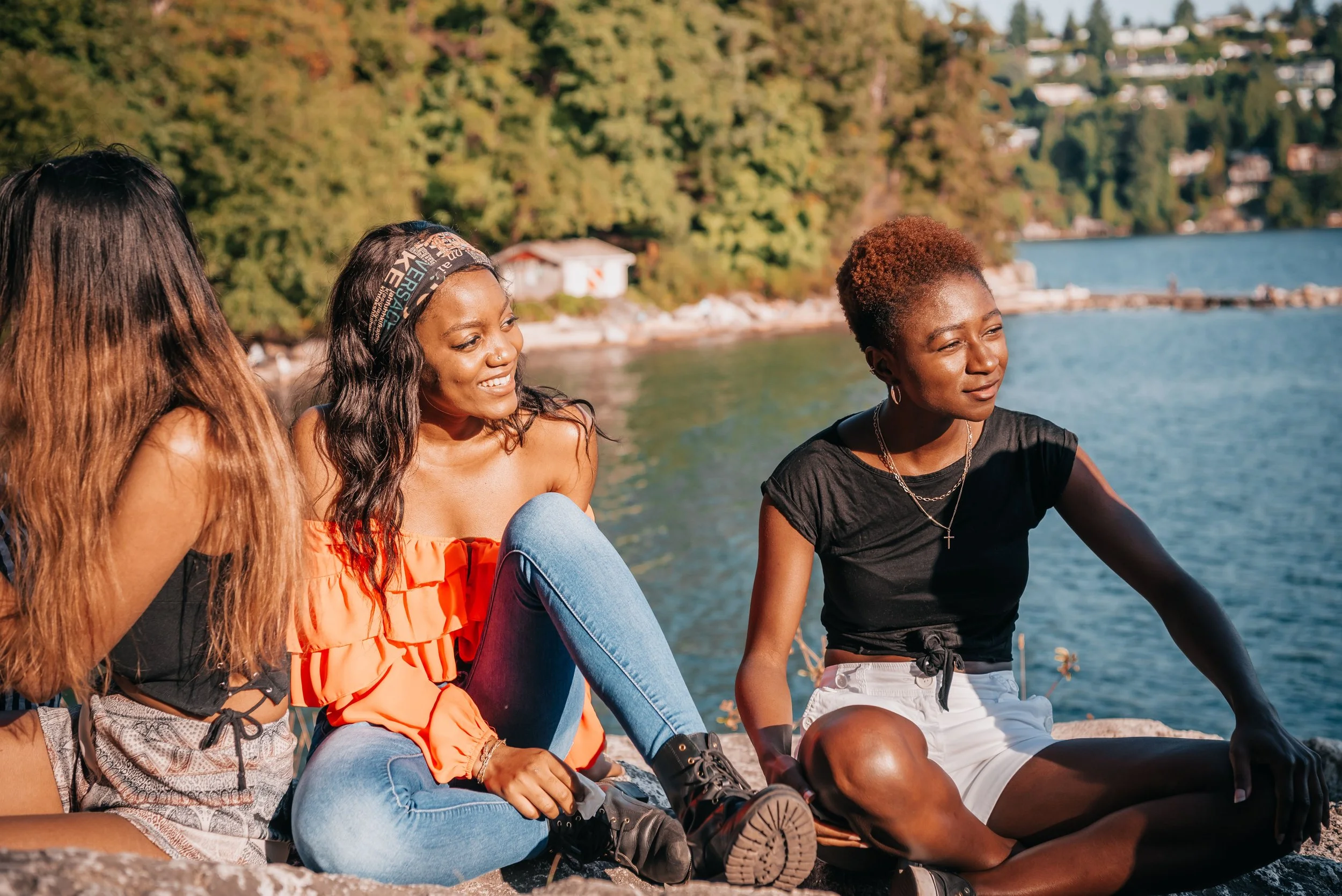 Four young women sitting by a body of water with trees and houses in the background, enjoying a sunny day.
