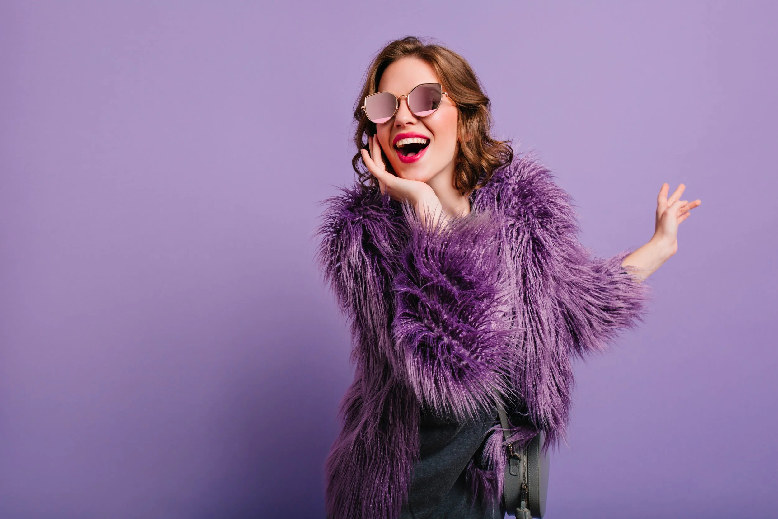 A smiling woman with brown hair wearing pink-tinted sunglasses, a purple furry coat, and a gray bag, posing against a purple background.