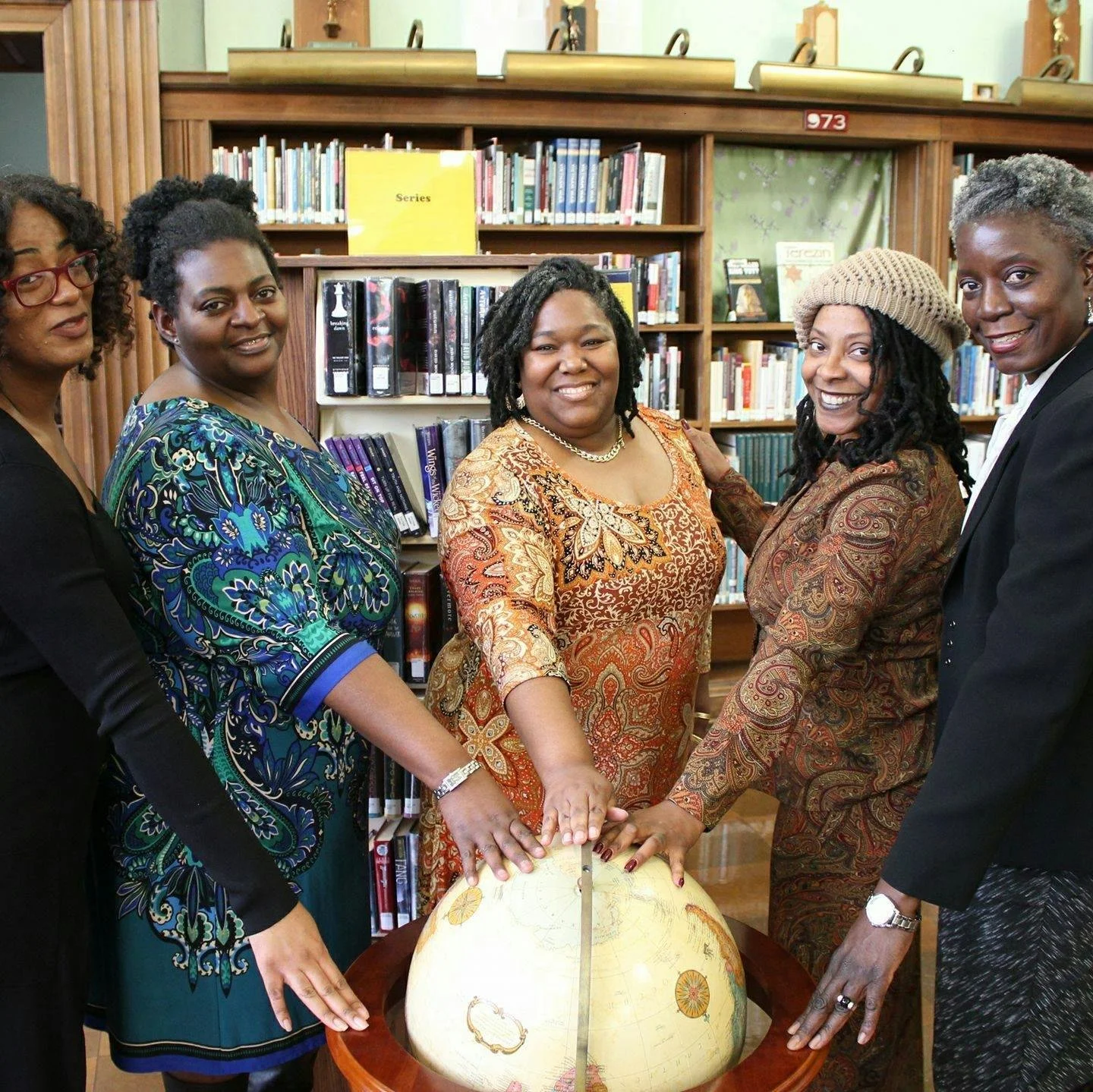 Five women standing in a library, holding hands over a globe placed on a table, all smiling and wearing colorful dresses.