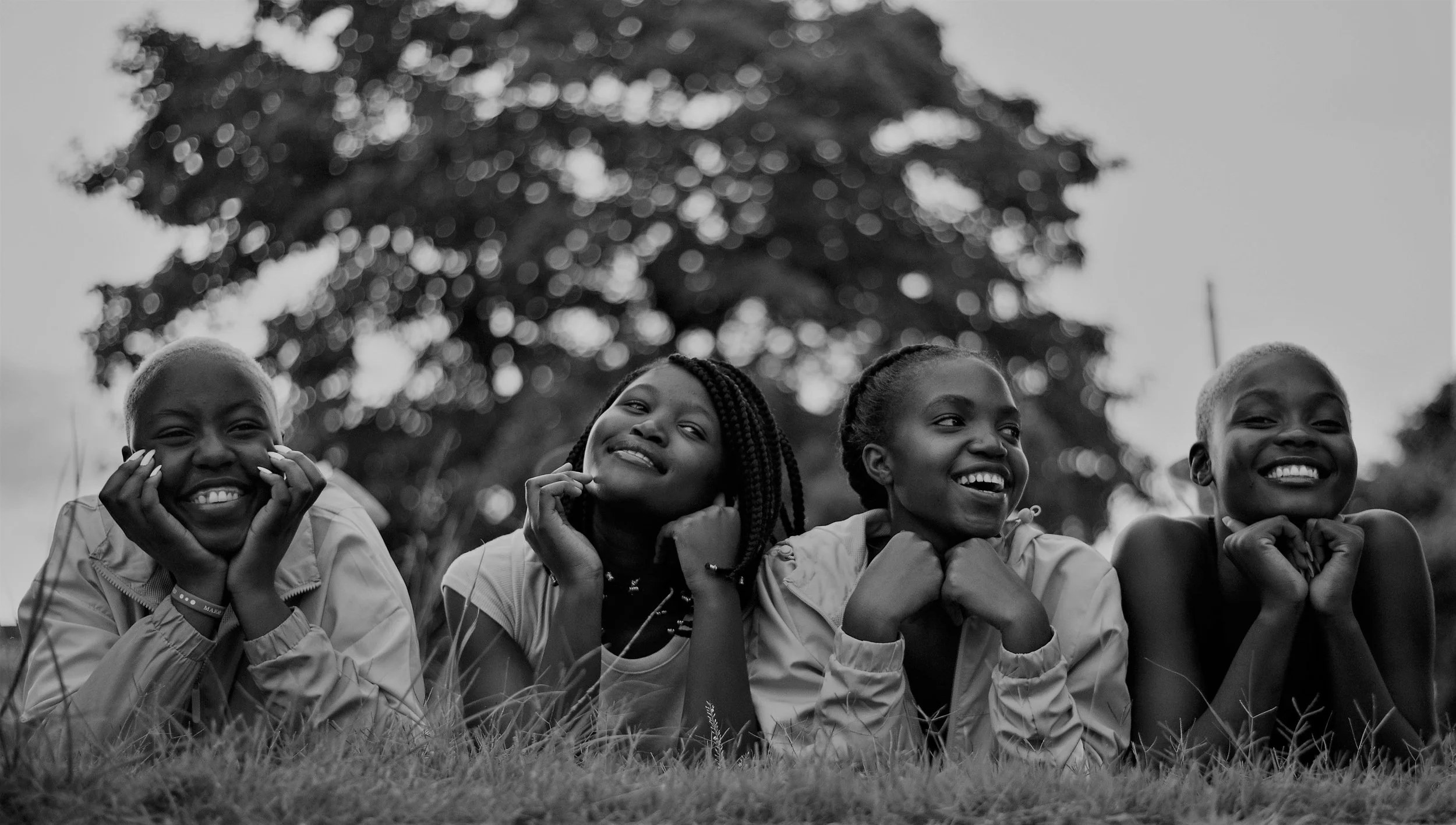 Four smiling children lying on the grass with a large tree in the background, captured in black and white.