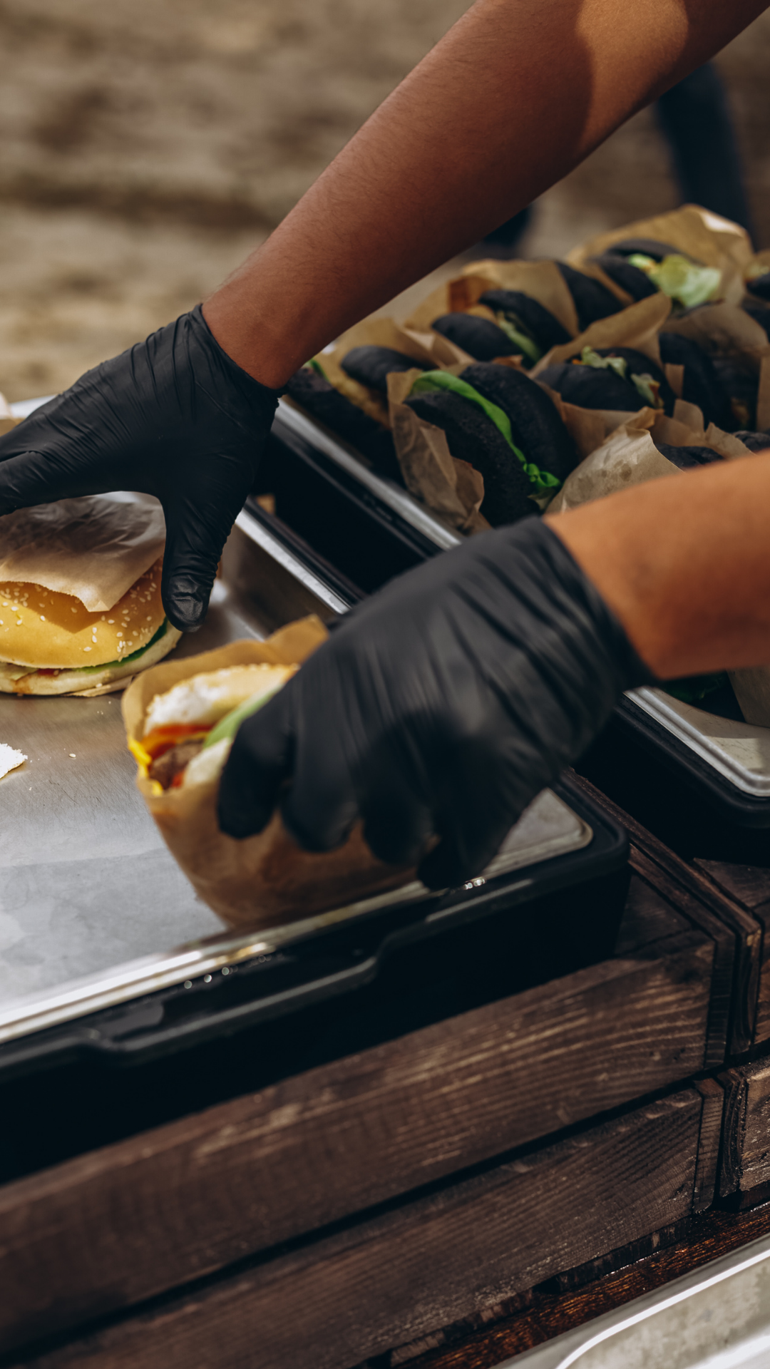 Personnes portant des gants noirs qui manipulent des sandwiches à base de viande, salade et sauce, dans une scène de préparation de nourriture.