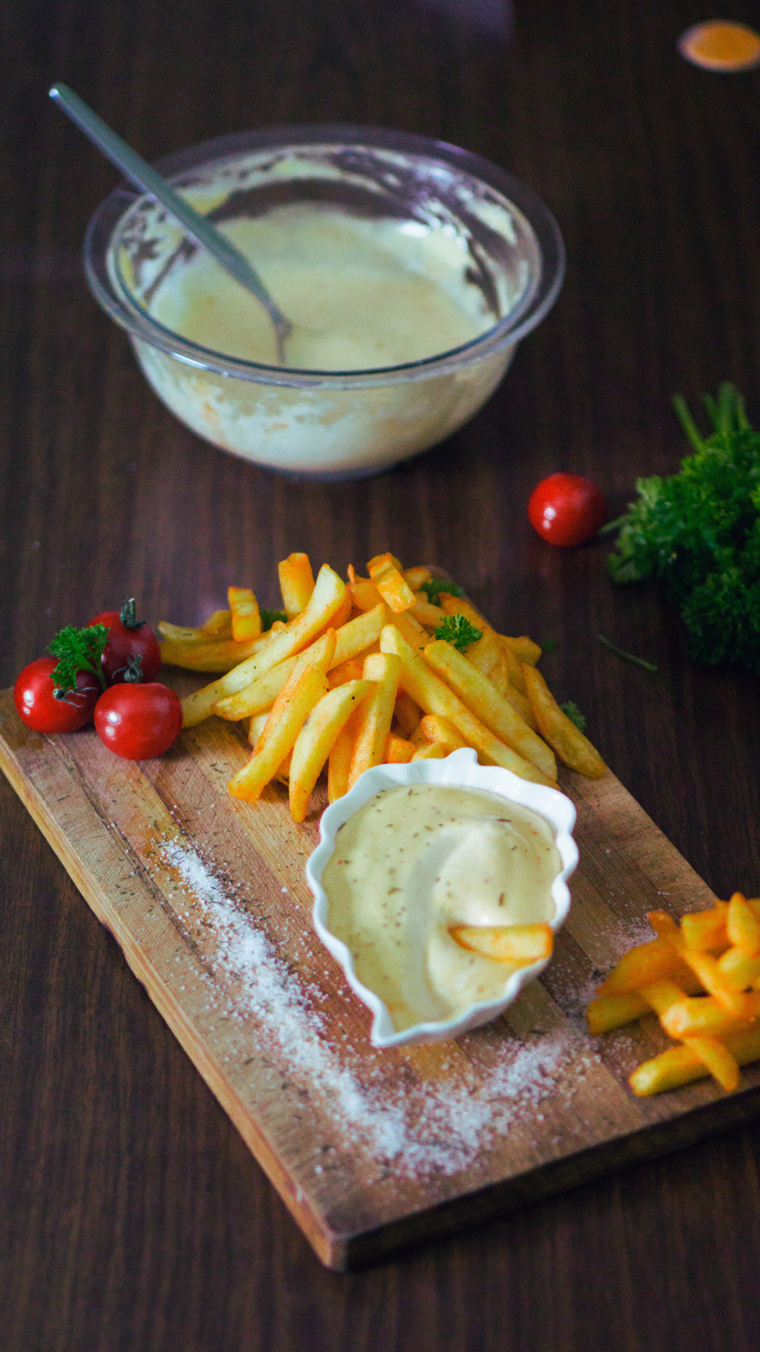 Frites avec sauce, tomates cerises, et herbes sur une planche en bois, devant un bol de mayonnaise, sur une table en bois.