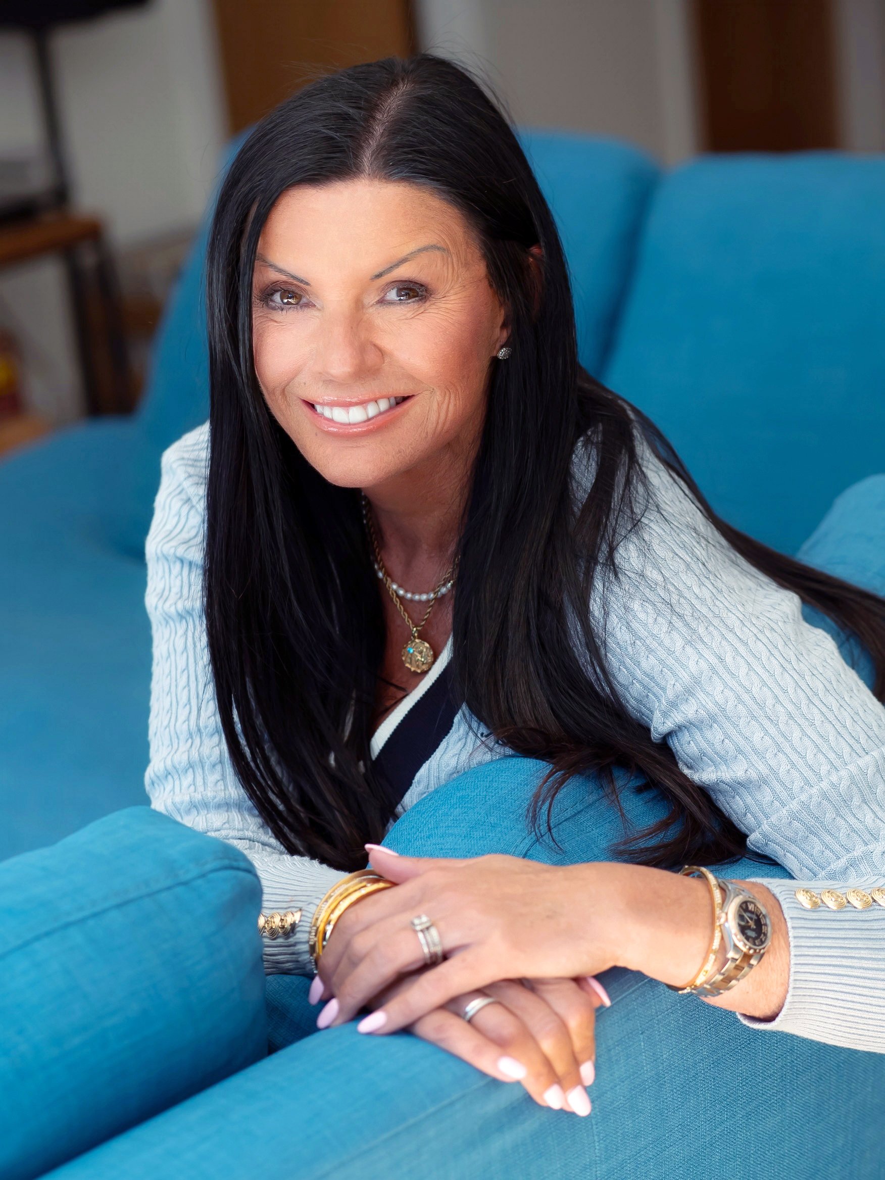 A woman with long black hair smiling, wearing jewelry, including a watch and rings, sitting on a turquoise couch with a blurred background.