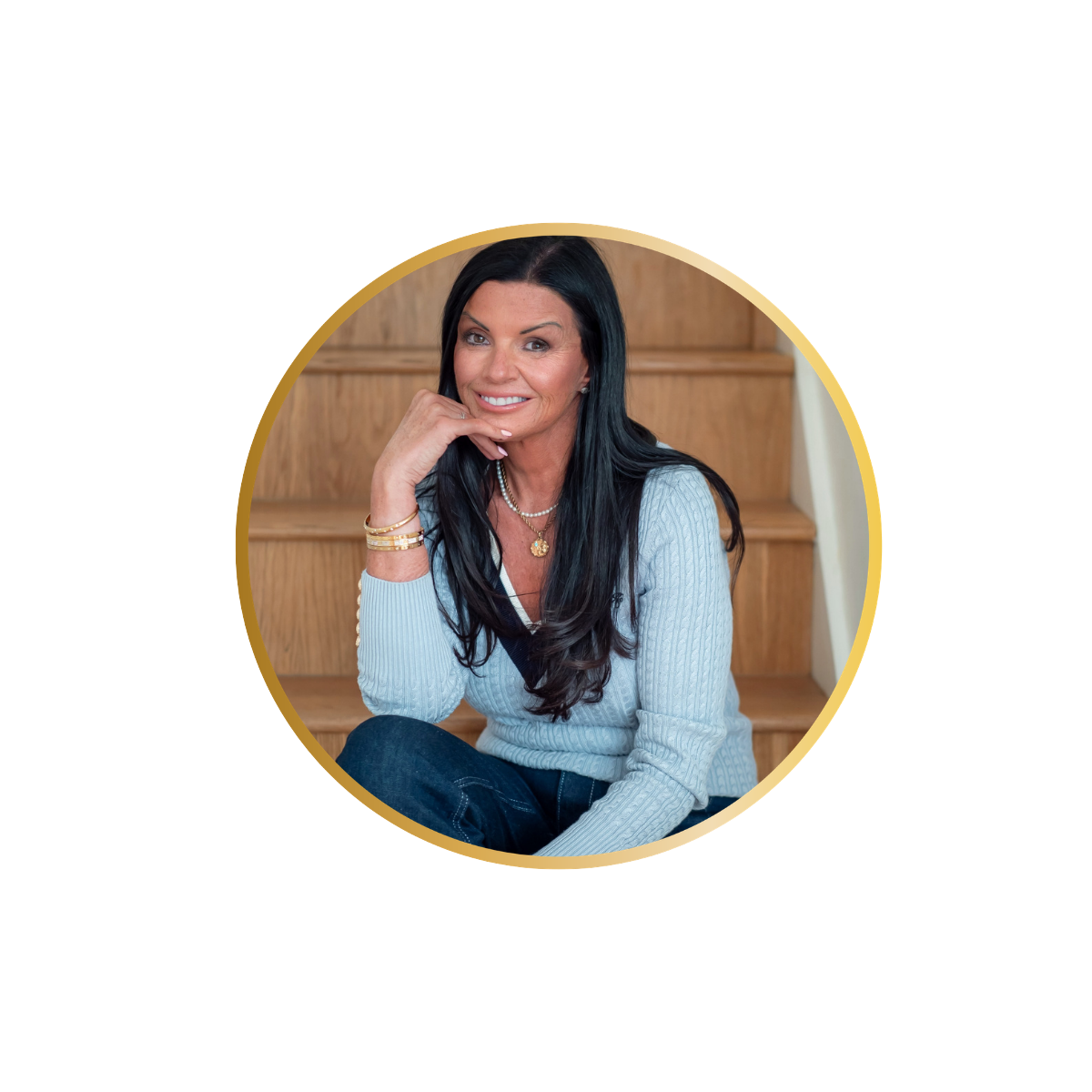 A woman with long black hair and light skin sitting on wooden stairs, wearing a light blue sweater, dark jeans, gold jewelry, and smiling at the camera.