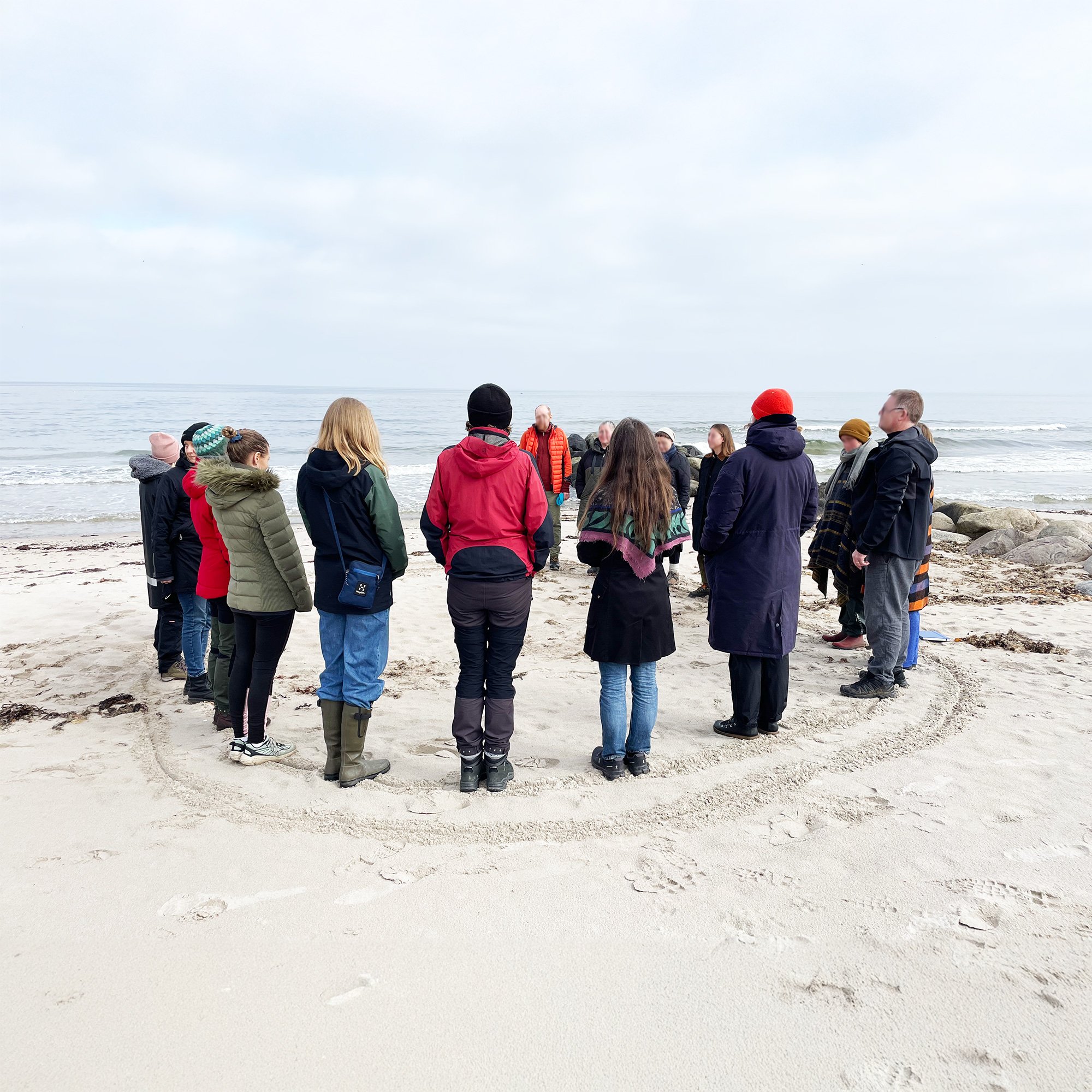 En gruppe mennesker står på en sandstrand med oceanet i baggrunden, nogle er iført jakker og hatte, hvilket indikerer køligt vejr.