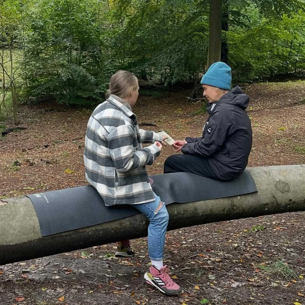To personer sidder på en træstamme i skoven. Den ene person modtager noget, muligvis penge eller et kort, fra den anden. Den ene person er klædt i en grå-hvid ternet jakke og lyse blå jeans, den anden i en sort jakke og blå hue.