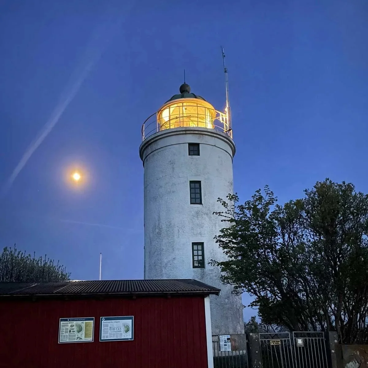 Billedet viser en lygtetårn om aftenen med fuldmånen i baggrunden og en klar blå himmel. Der er nogle træer og en bygning med informationsskilte foran tårnet.