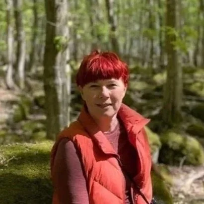 En kvinde med rødt hår smiler udendørs, holder en bog om bæredygtighed, bærer en brunt uldtrøje og en lang brun nederdel, med solbriller på hovedet og en kamera hængende om halsen, omgivet af grønne træer og blå himmel. Anna Taws, Reflektionsrum.