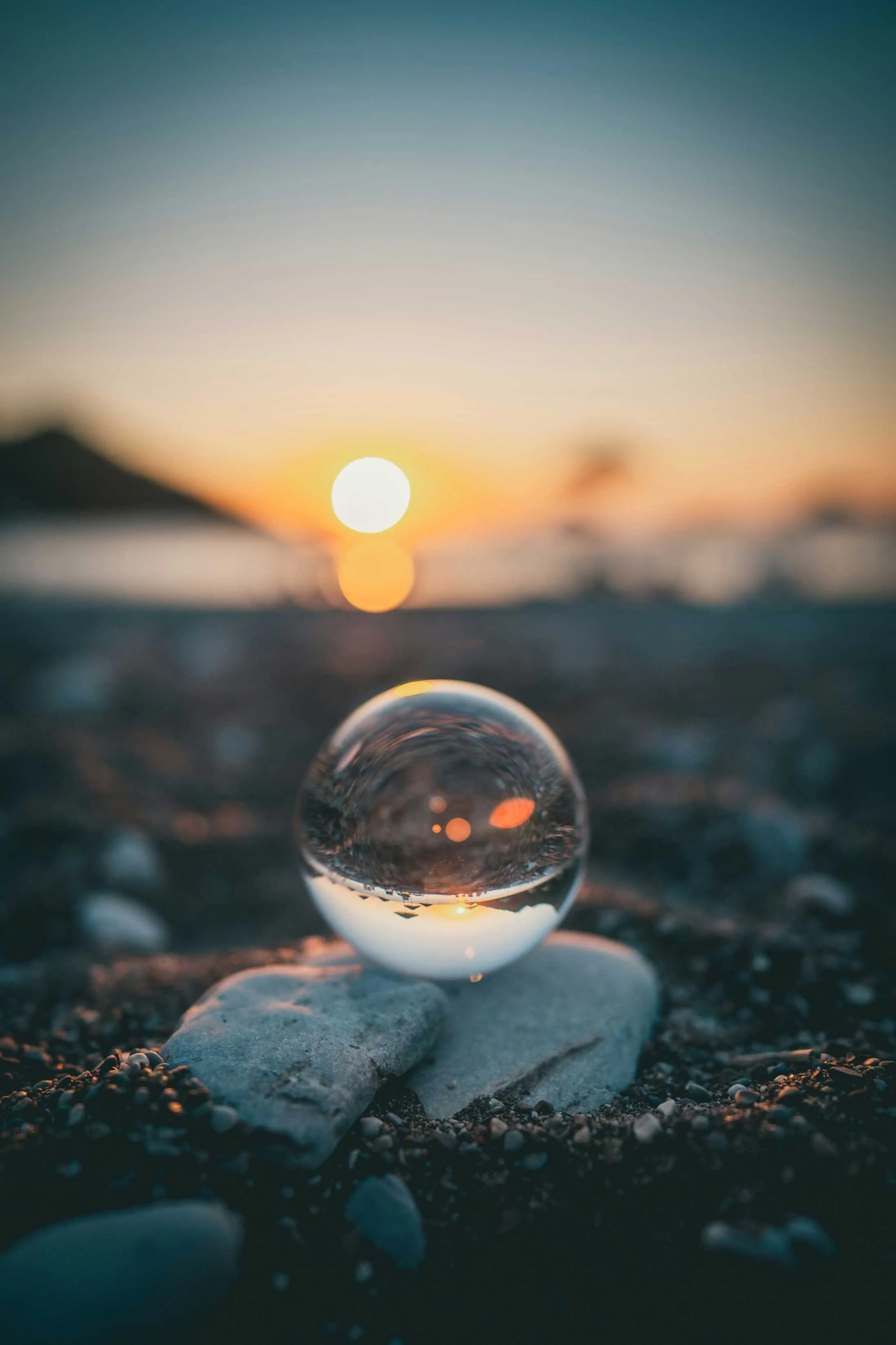 En gennemsigtig glasbold placeret på en sandet strand med småsten, der reflekterer solnedgangen i horisonten, som er tydeligt synlig i baggrunden.