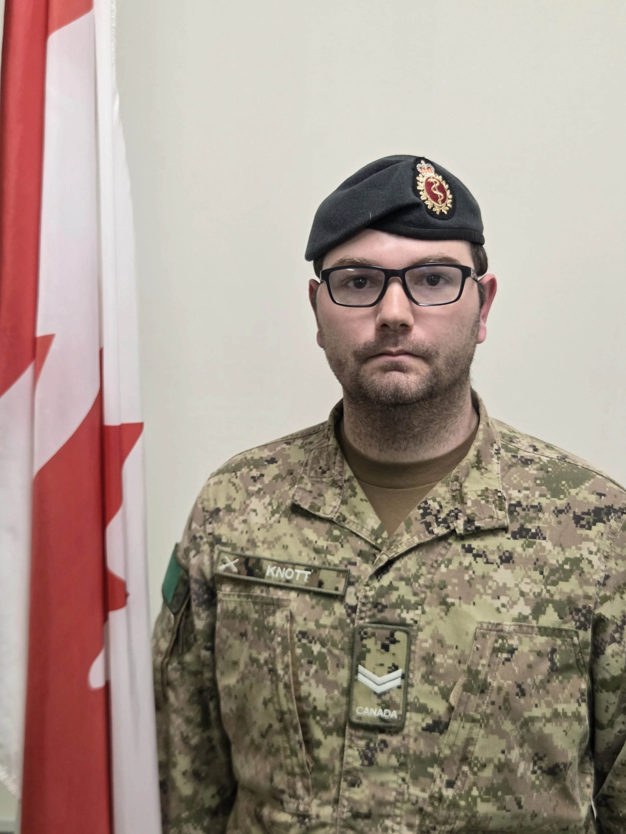 A man in military uniform, wearing glasses and a beret with a patch, standing in front of a beige wall and partial view of a Canadian flag.