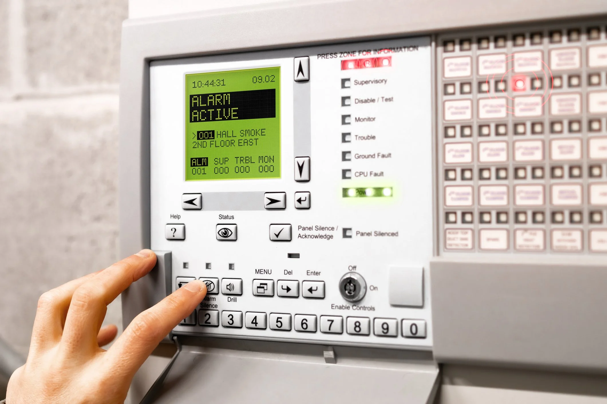 A person using a fire alarm control panel with a digital display indicating the alarm is active, and several buttons and indicator lights on the panel.
