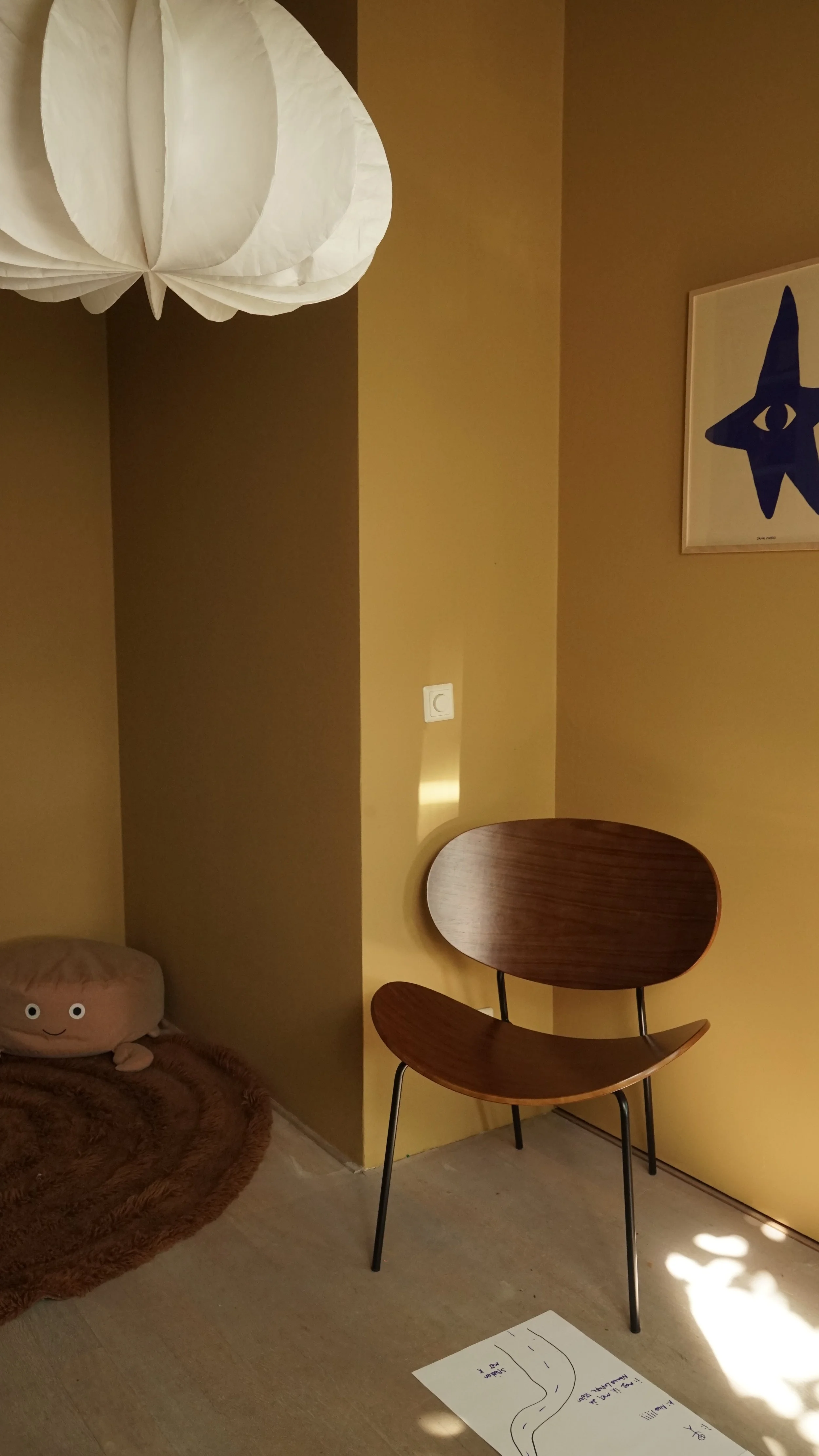 Binnenruimtelijke setting met gele wand, houten stoel, hanglamp en kunstwerk met een ster op de muur. Op de vloer ligt een handgeschreven diagram.