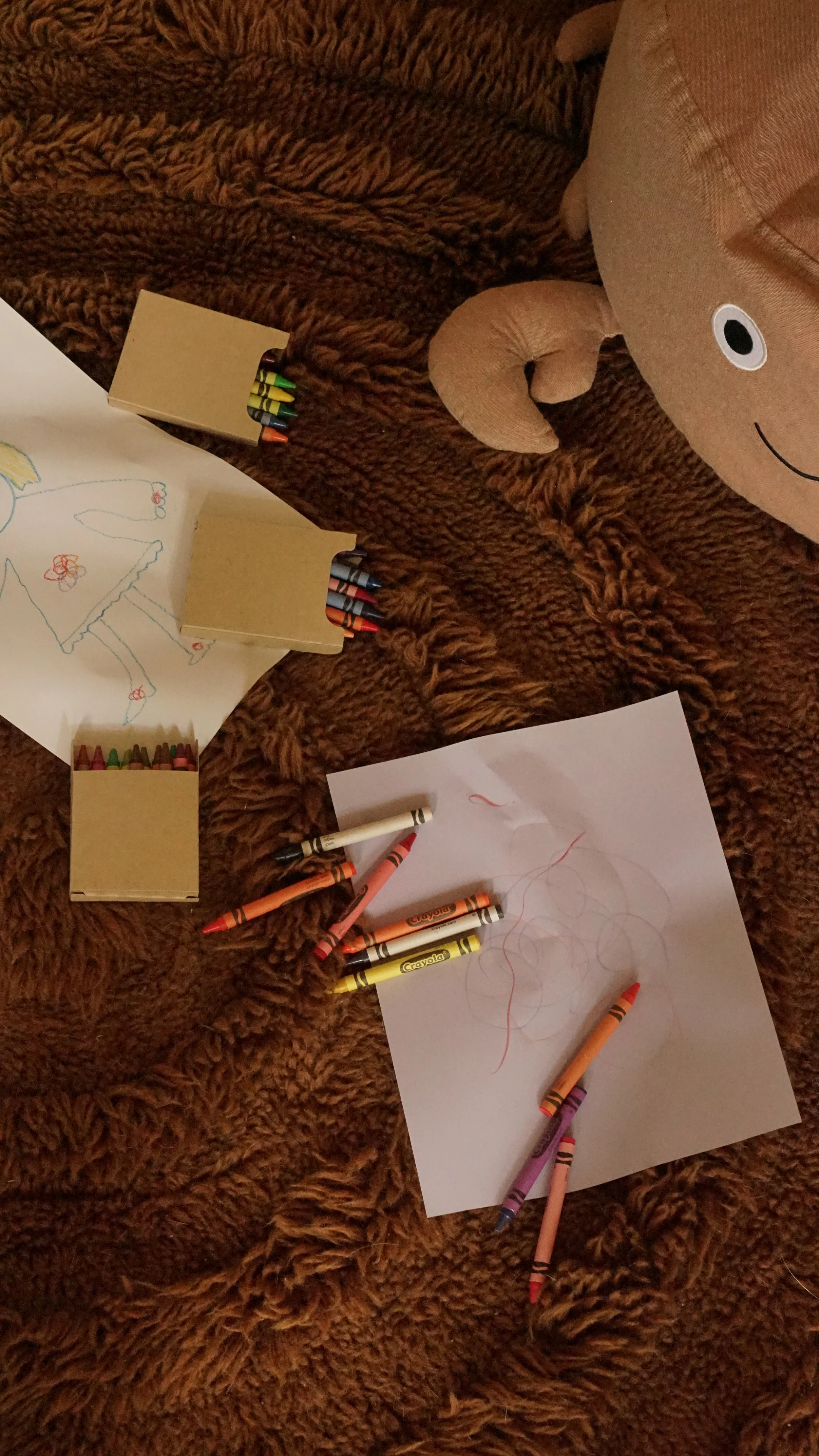 Op een bruine, zachte kleed liggen schetsen, gekleurde waskrijtjes en een groot teddybeer.