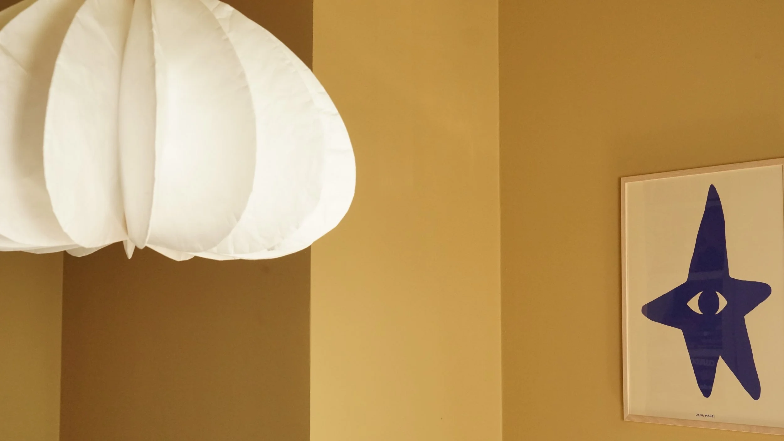 Een witte hanglamp gezien van onderaf, naast een gele muur en een kunstwerk met een blauwe ster en een oog. Onder het kunstwerk staat de naam Jana Marem.