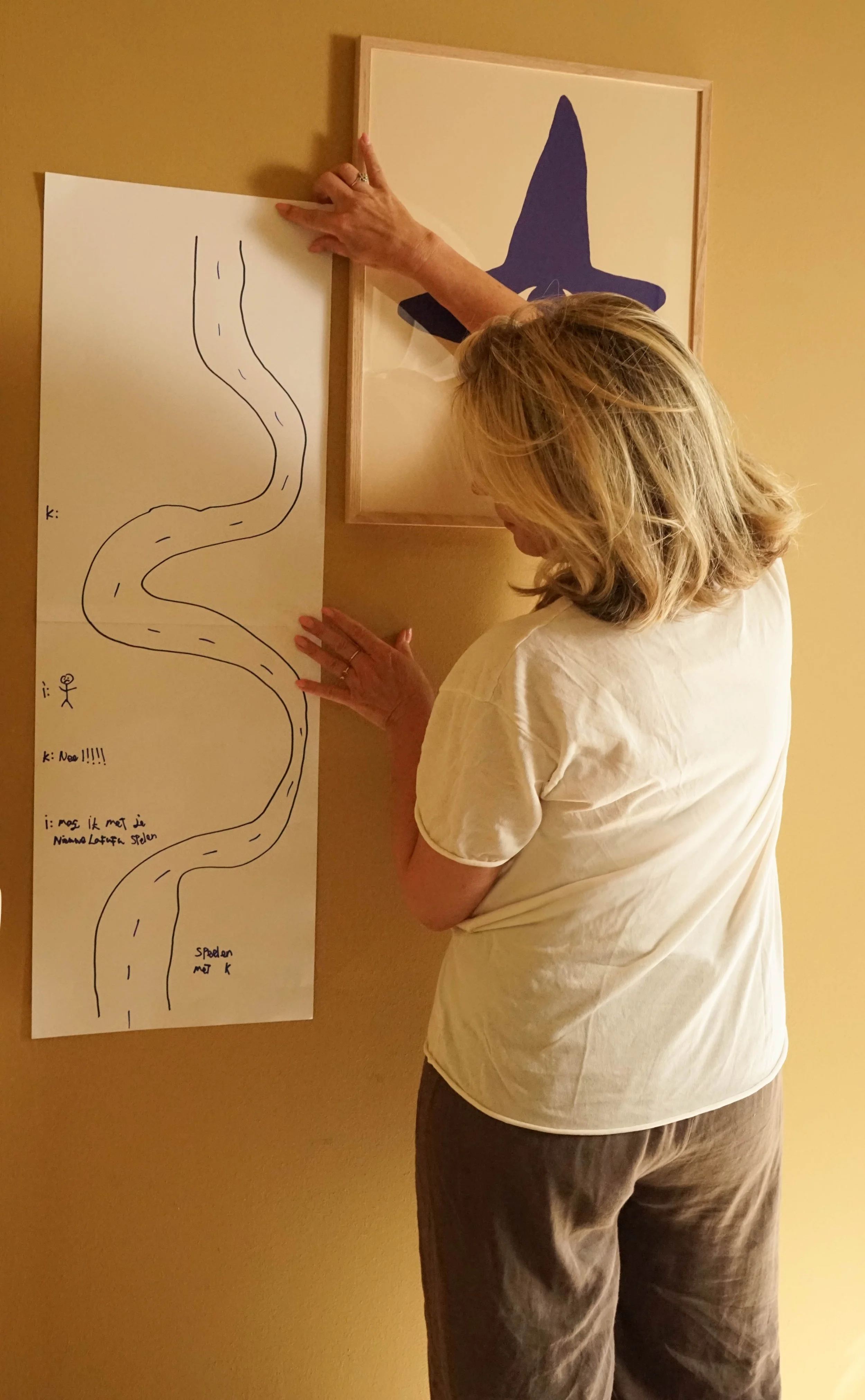 Een vrouw die een lange scheurlijn tekent op een groot vel papier dat aan de muur is bevestigd. Ze wijst naar het papier met haar rechterhand en heeft haar hoofd naar beneden gericht, haar ogen lijken op het papier gericht. Op het papier staan instructies in het Nederlands.