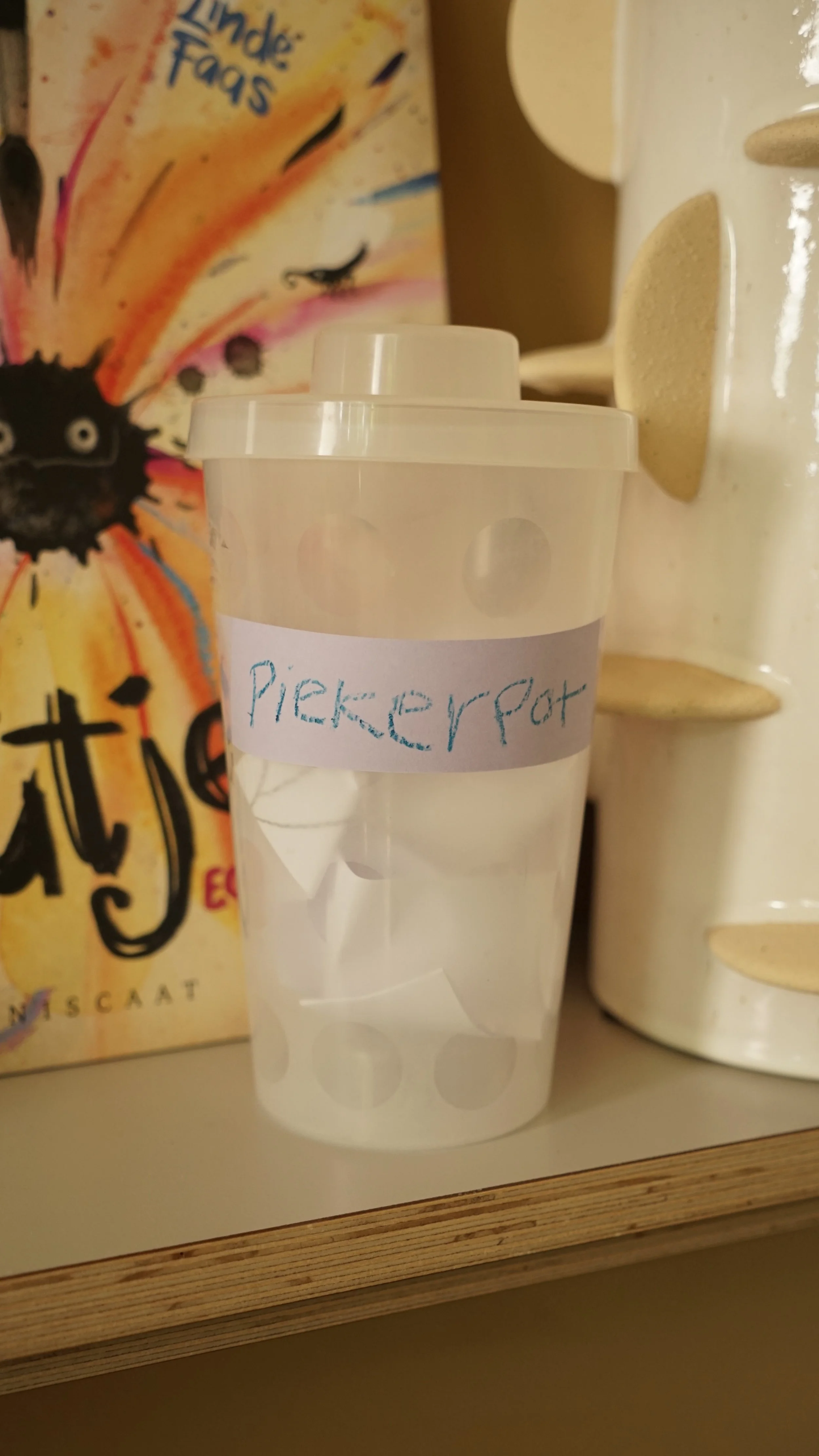 Een doorzichtig plastic potje met een wit label dat 'Pickerpot' schrijft. Het potje staat op een plank tussen een doos met kleurrijk ontwerp en een wit houdbaarhedenrek.
