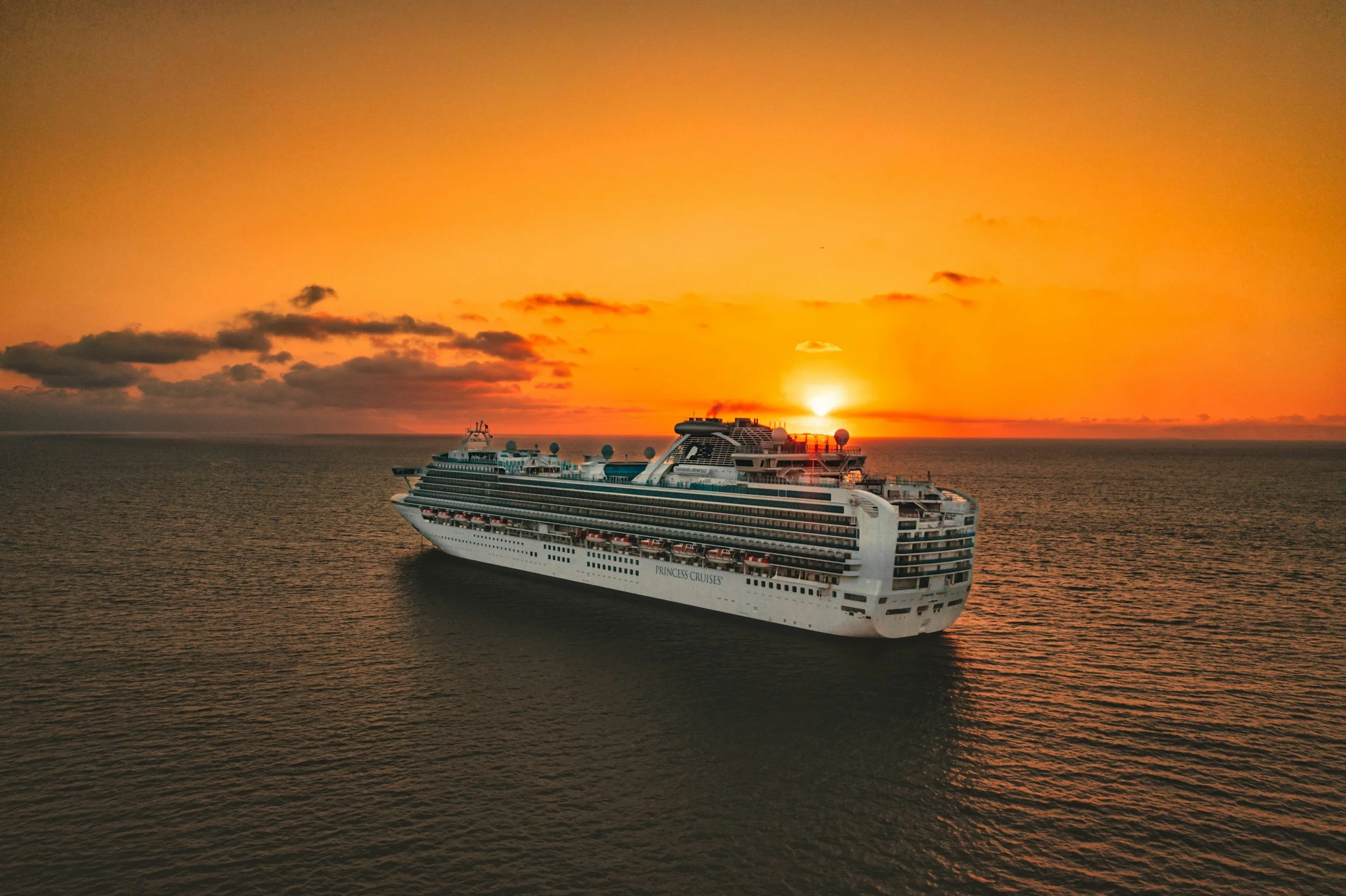 A large white cruise ship labeled 'Princess Cruises' sailing on the ocean during sunset with orange sky and some clouds in the background.