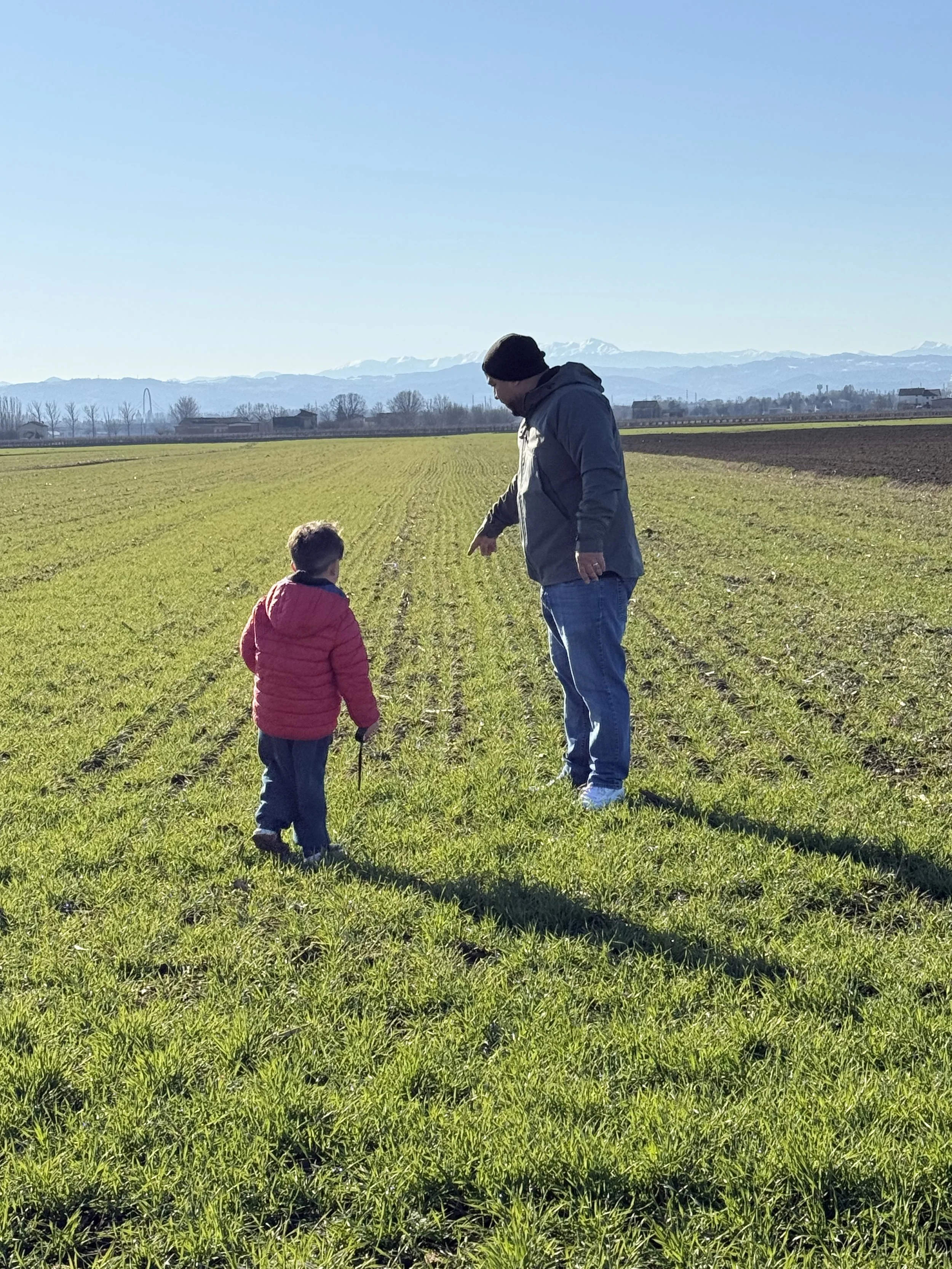 An adult man in a gray hoodie and beanie talking to a young boy in a red jacket in a green, open field with mountains in the distance and clear blue sky.