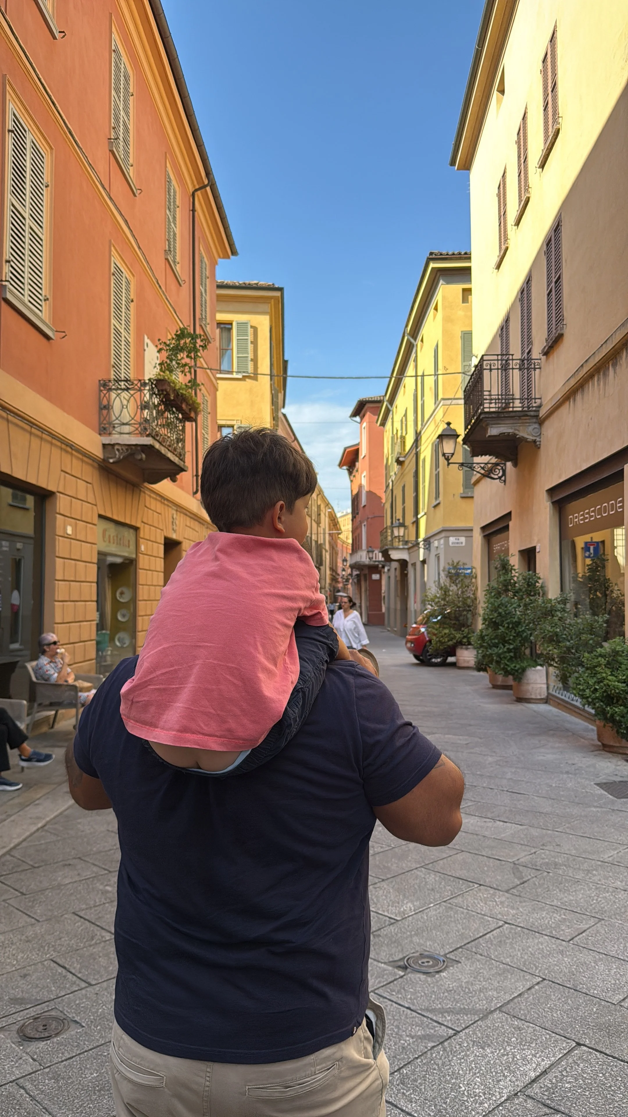 A person carrying a child on their shoulders walking down a colorful European street with pastel-colored buildings, balconies, and a clear blue sky.