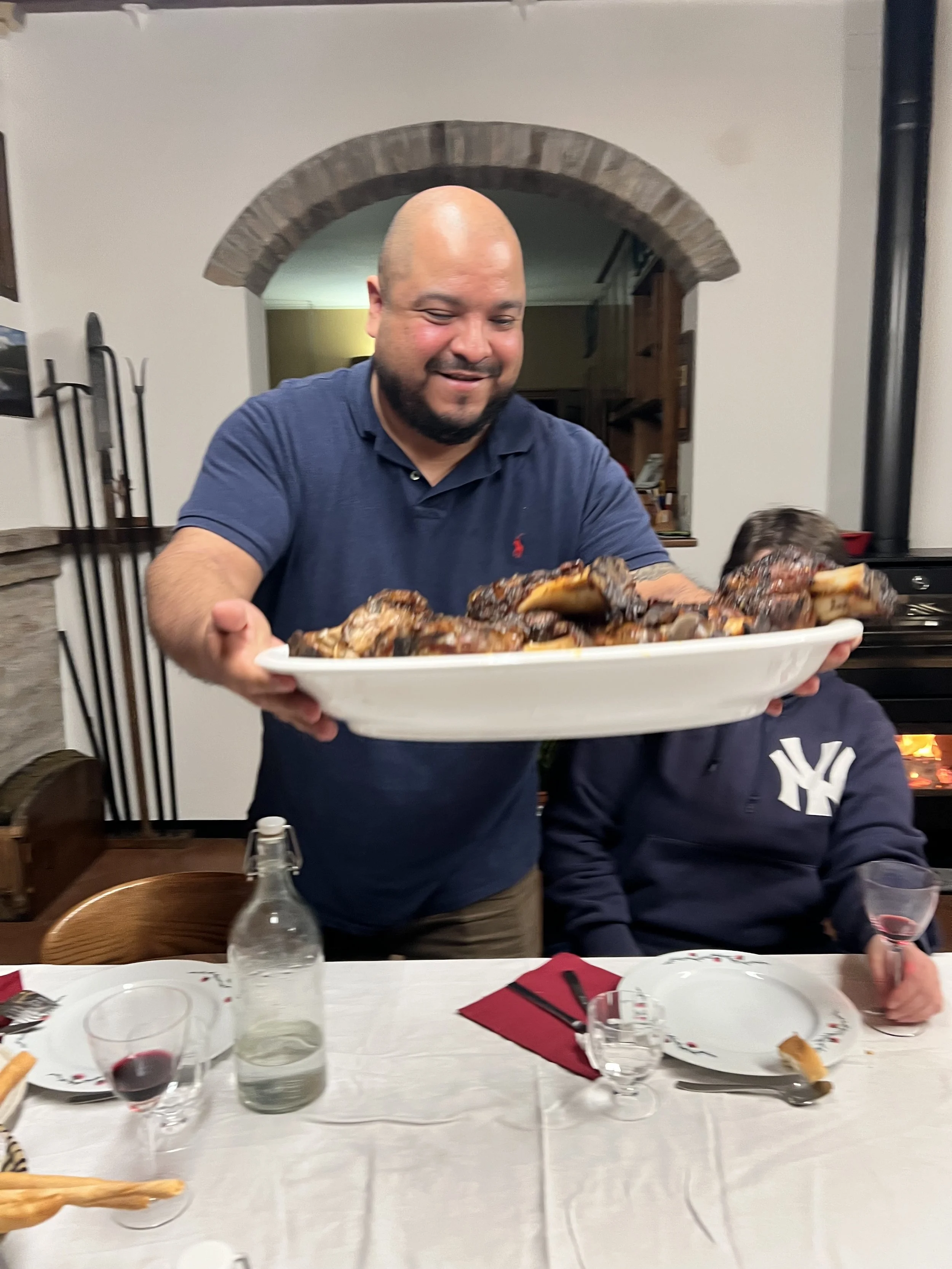 A man with a bald head and beard smiling while holding a large platter of cooked meat at a family dinner. A boy wearing a navy hoodie with a white logo is sitting beside him. The table is set with glasses, plates, a water bottle, and a red napkin. In the background, there is a brick archway and a cozy home interior.