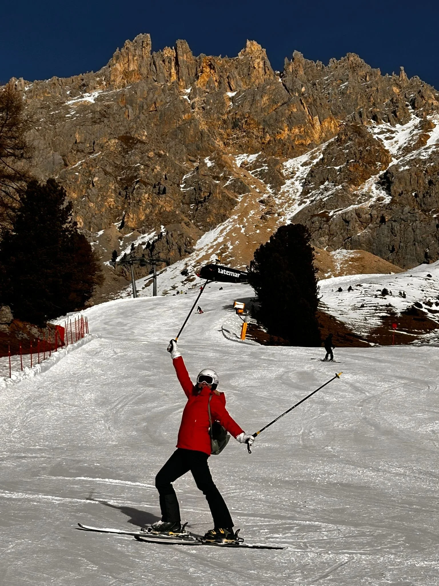 A person in a red jacket and black pants on skis, raising their arm in a celebratory gesture, on a snowy ski slope. There are other skiers in the background, surrounded by tall trees and a mountain with rocky cliffs and patches of snow, under a clear dark blue sky.