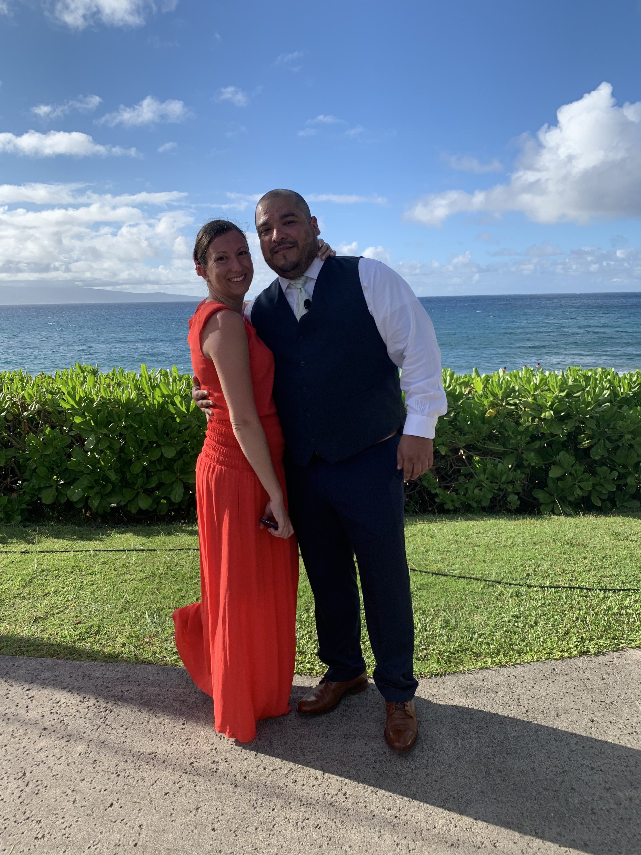 A couple is posing together outdoors near the ocean, with clear blue sky and scattered clouds. The woman is wearing a red sleeveless dress, and the man is dressed in a white shirt, dark vest, and dark pants. They are smiling and standing close to each other, with green bushes behind them and the ocean in the background.