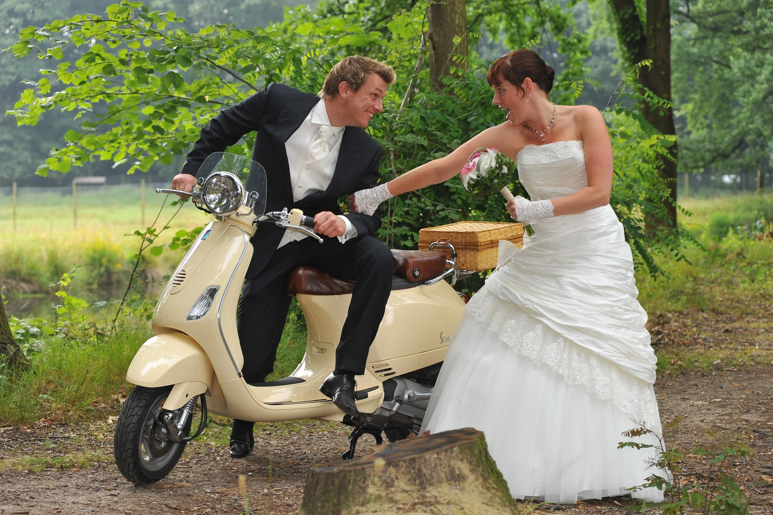 Een bruid in een witte trouwjurk en een bruidegom in een zwart pak op een scooter in een bosrijke omgeving, ze lachen naar elkaar.
