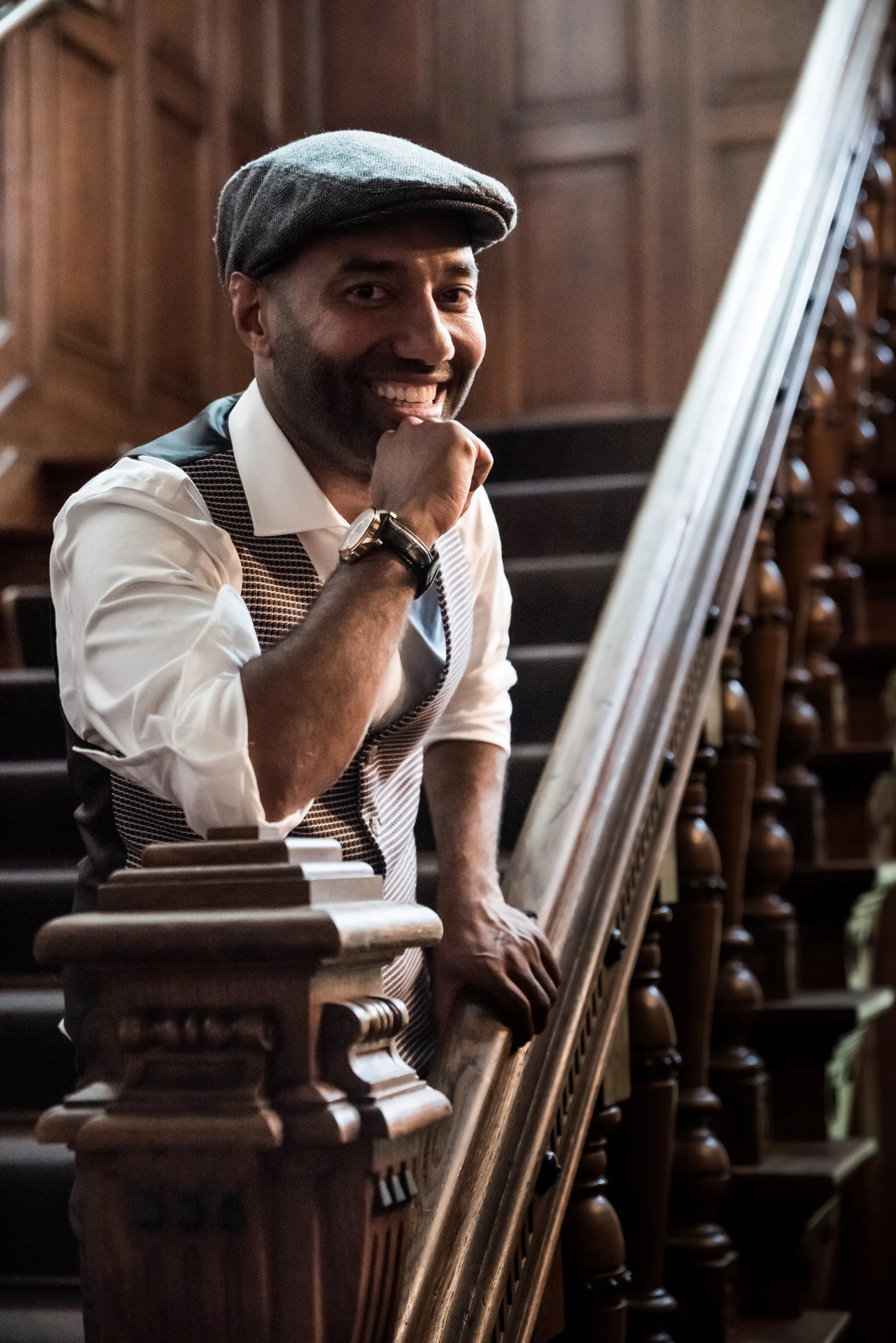 Een man glimlacht in een oude kerk of kathedraal, terwijl hij leunt op een houten balustrade met een hand onder zijn kin. Hij draagt een grijze pet, een witte overhemd, en een horloge.