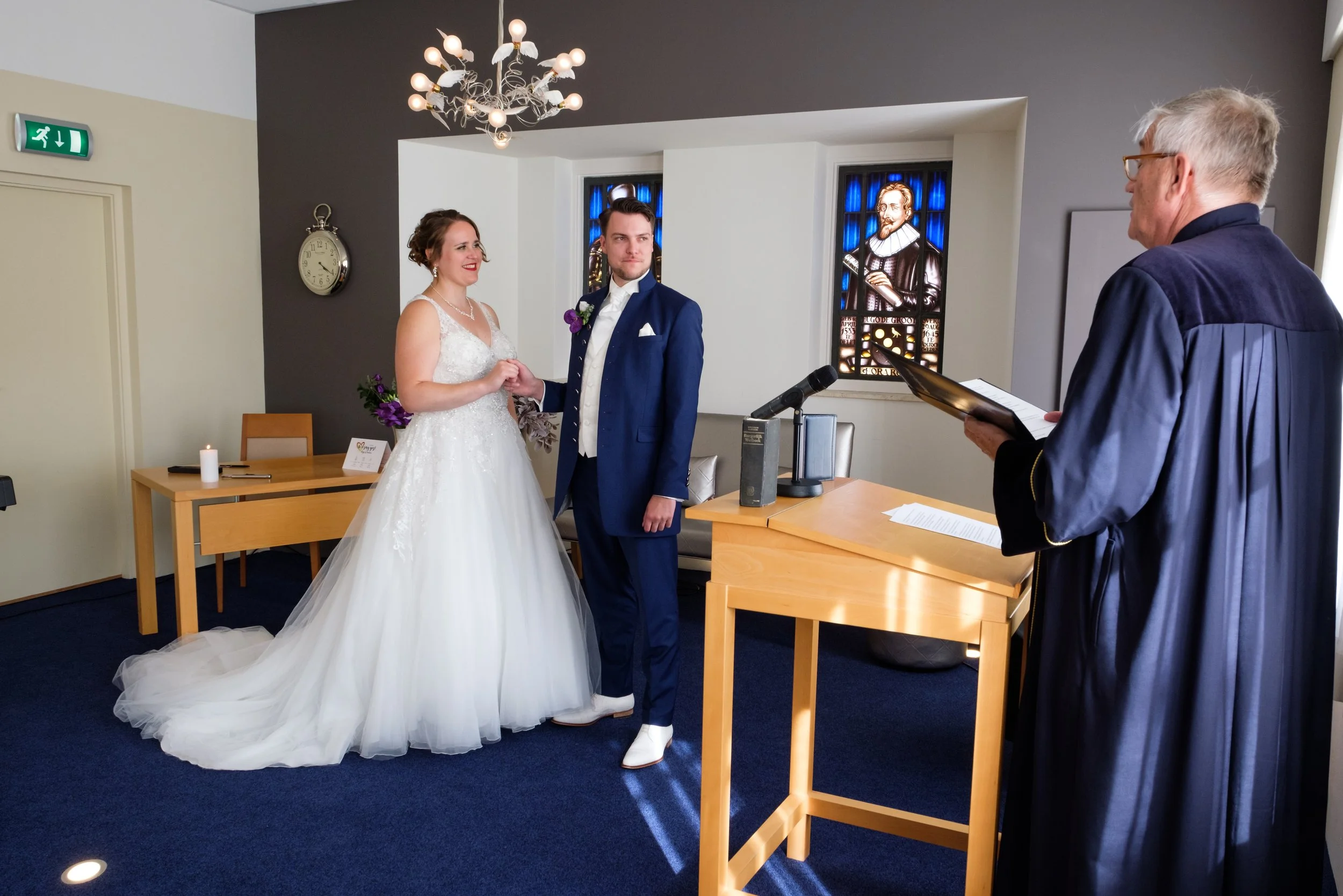 Een bruid en bruidegom tijdens hun huwelijksgeloof, terwijl een officier hen zal huwen, in een ruimte met twee gebrandschilderde ramen achter hen.