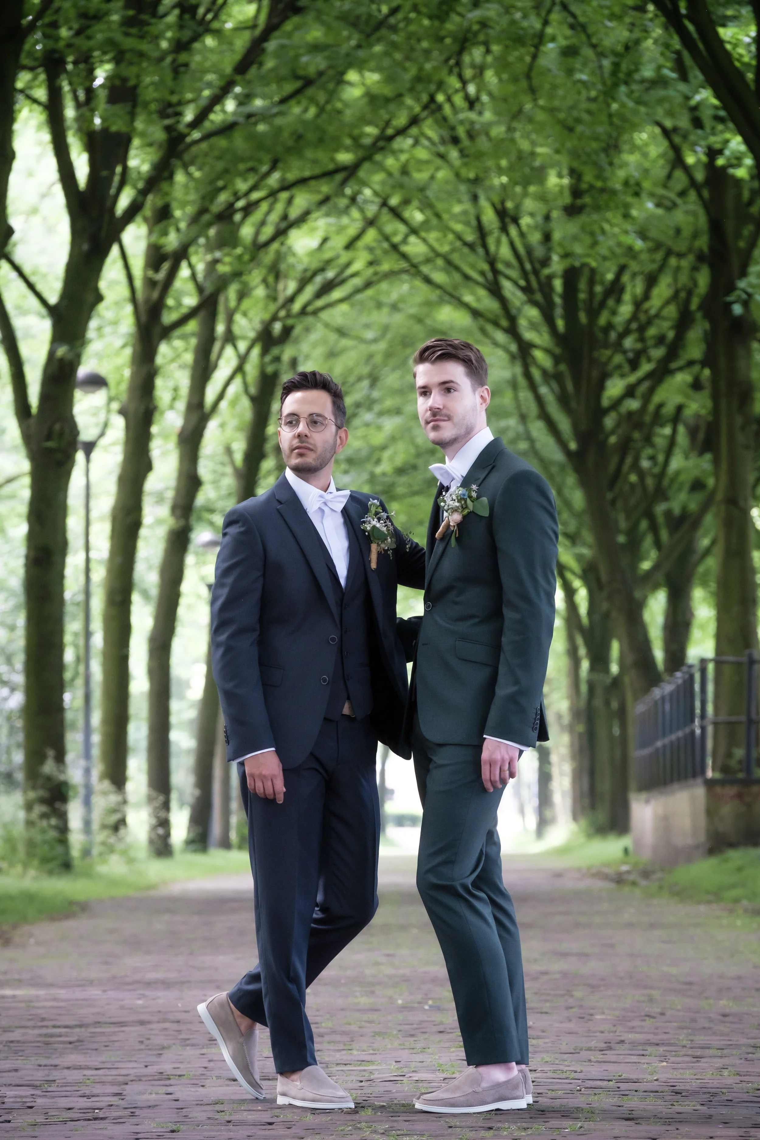 Twee mannen in formele pakken staan samen in een park, omringd door groene bomen.