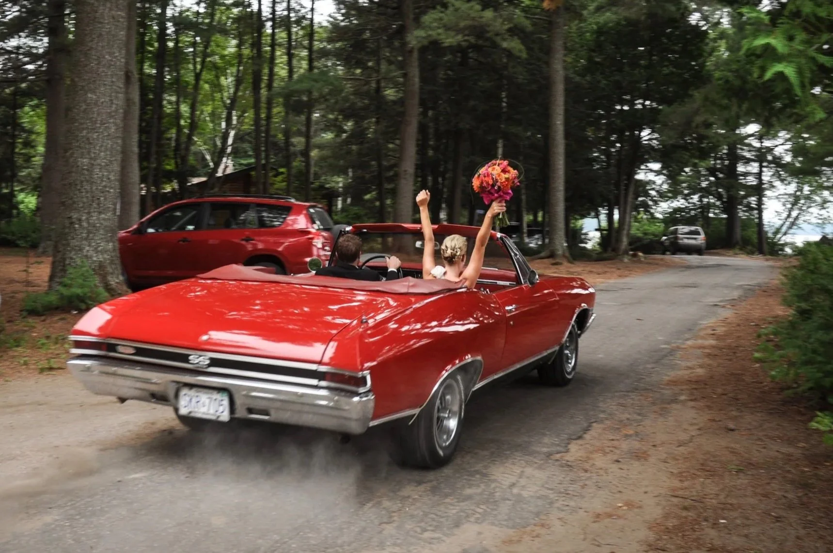 Een bruid en bruidegom in een rode klassieke cabriolet rijden door een bos, de bruid houdt een bloemenboeket omhoog en zwaait.