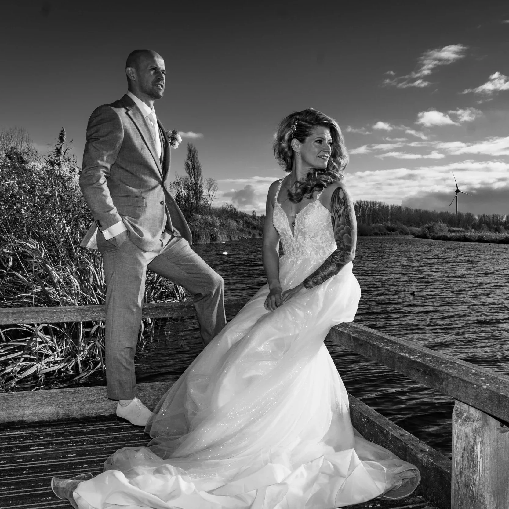 Zwart-wit foto van een koppel in trouwkleding, zittend op een houten steiger bij een rivier. De man draagt een grijze pak en de vrouw draagt een witte bruiloftsjurk met tattoos op haar armen. Op de achtergrond zijn bomen en een windmolen te zien onde