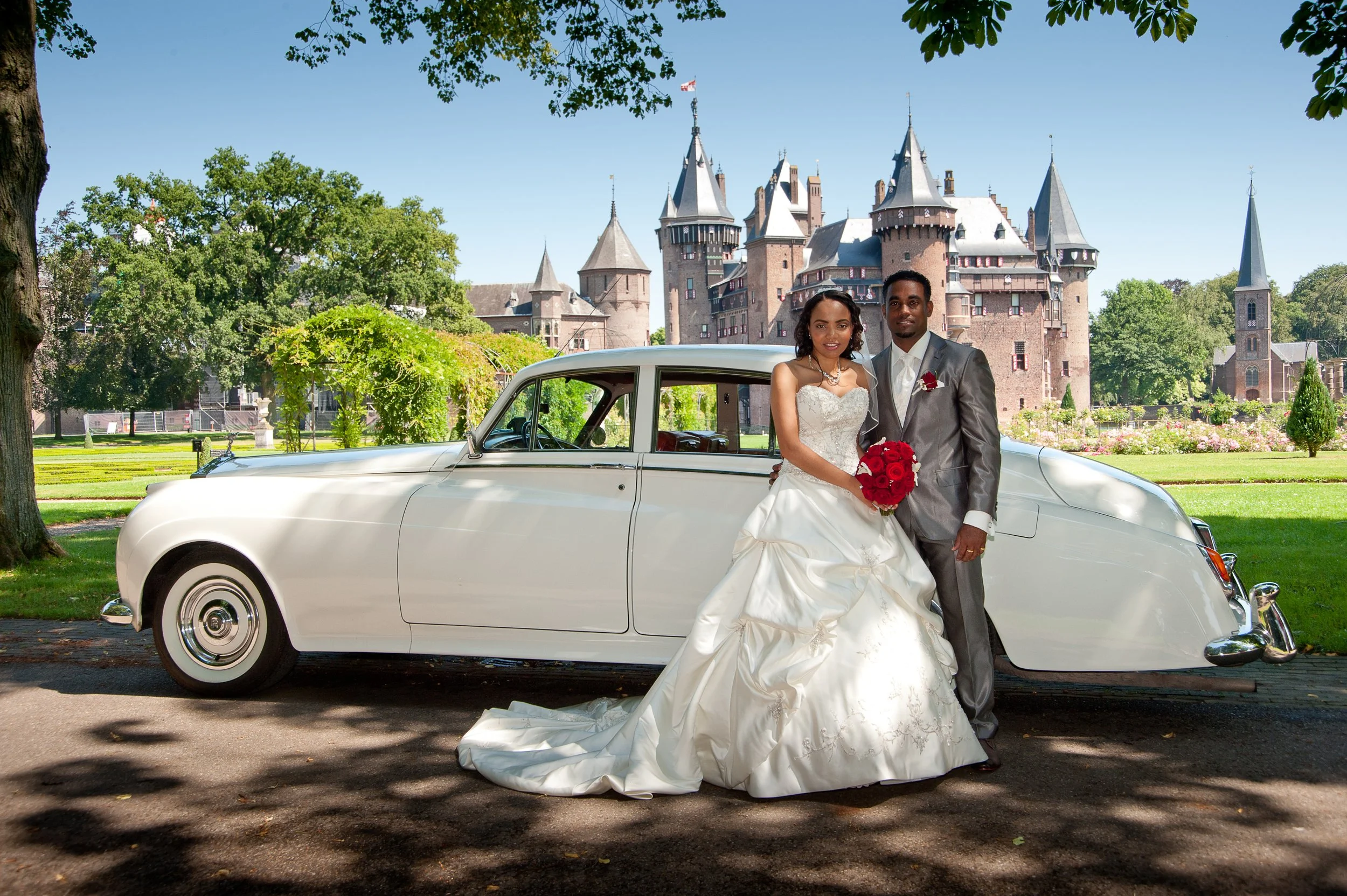 Een getrouwde man en vrouw in trouwjurk en pak staan voor een klassieke witte auto op een park met kasteel op de achtergrond.