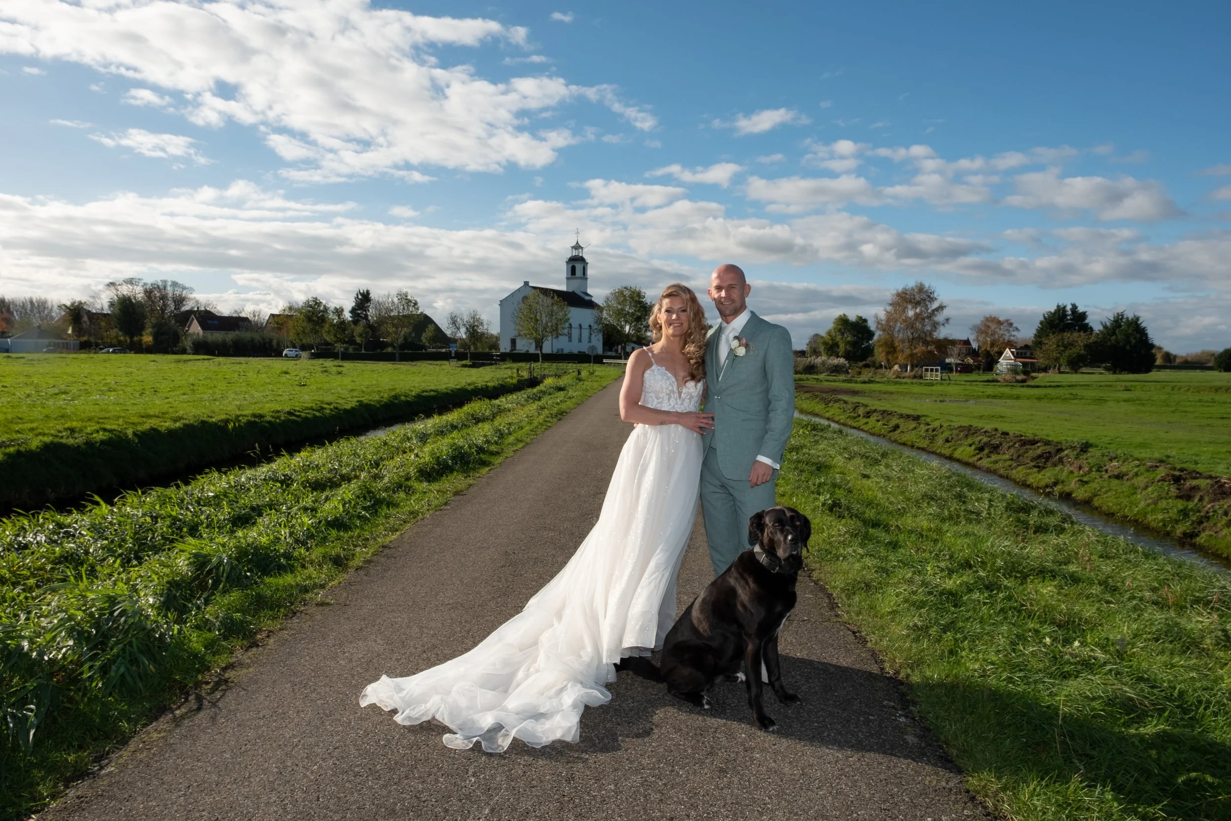 Een bruid en bruidegom staan samen op een smal pad in een landelijke omgeving, met een hond, onder een partly bewolkte blauwe lucht en een kerk op de achtergrond.