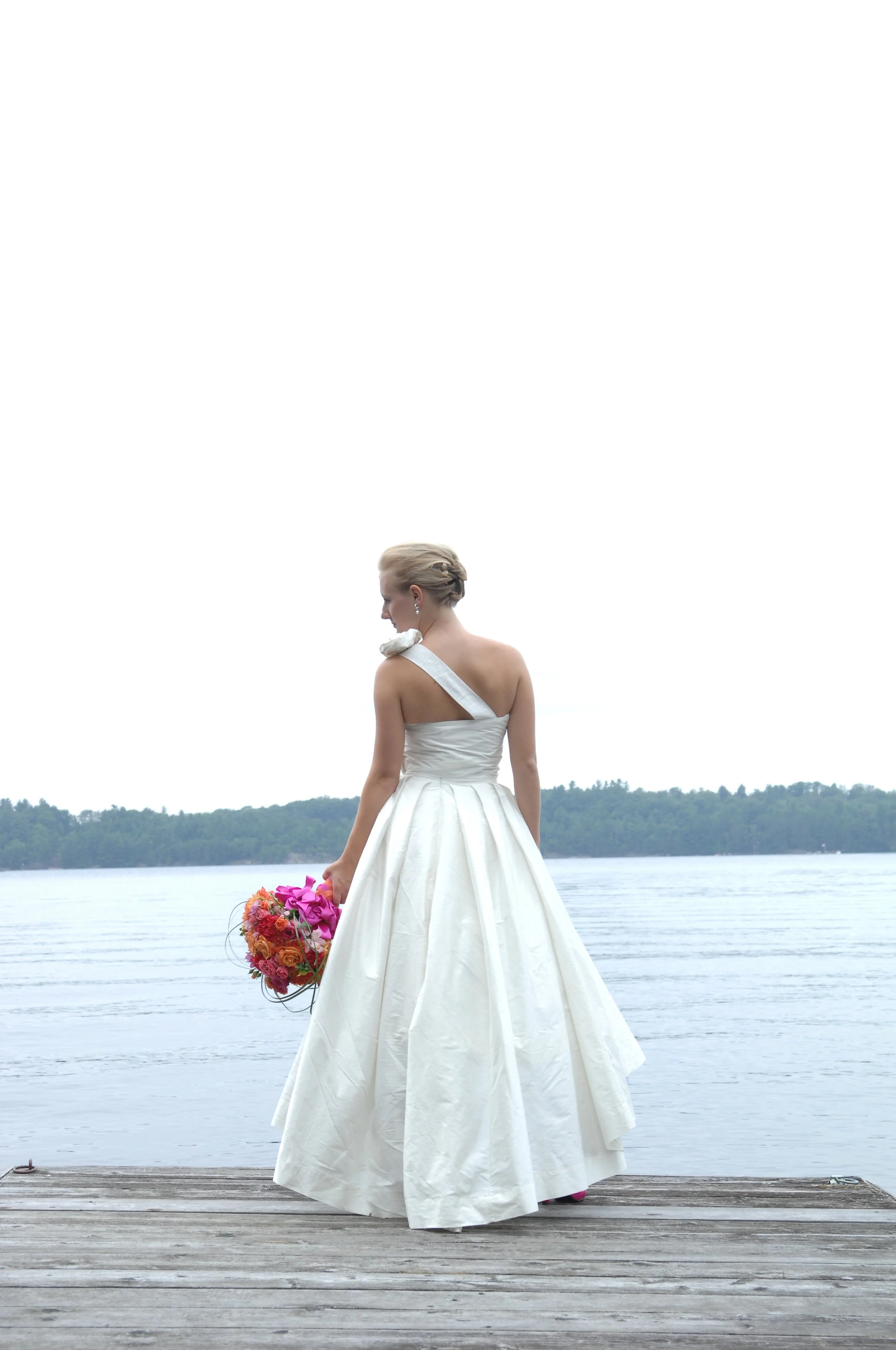 Een vrouw in een witte trouwjurk staat op een houten pier naast een water, met een bos bloemen in haar hand, en kijkt naar beneden. Op de achtergrond is het water en een bos te zien.