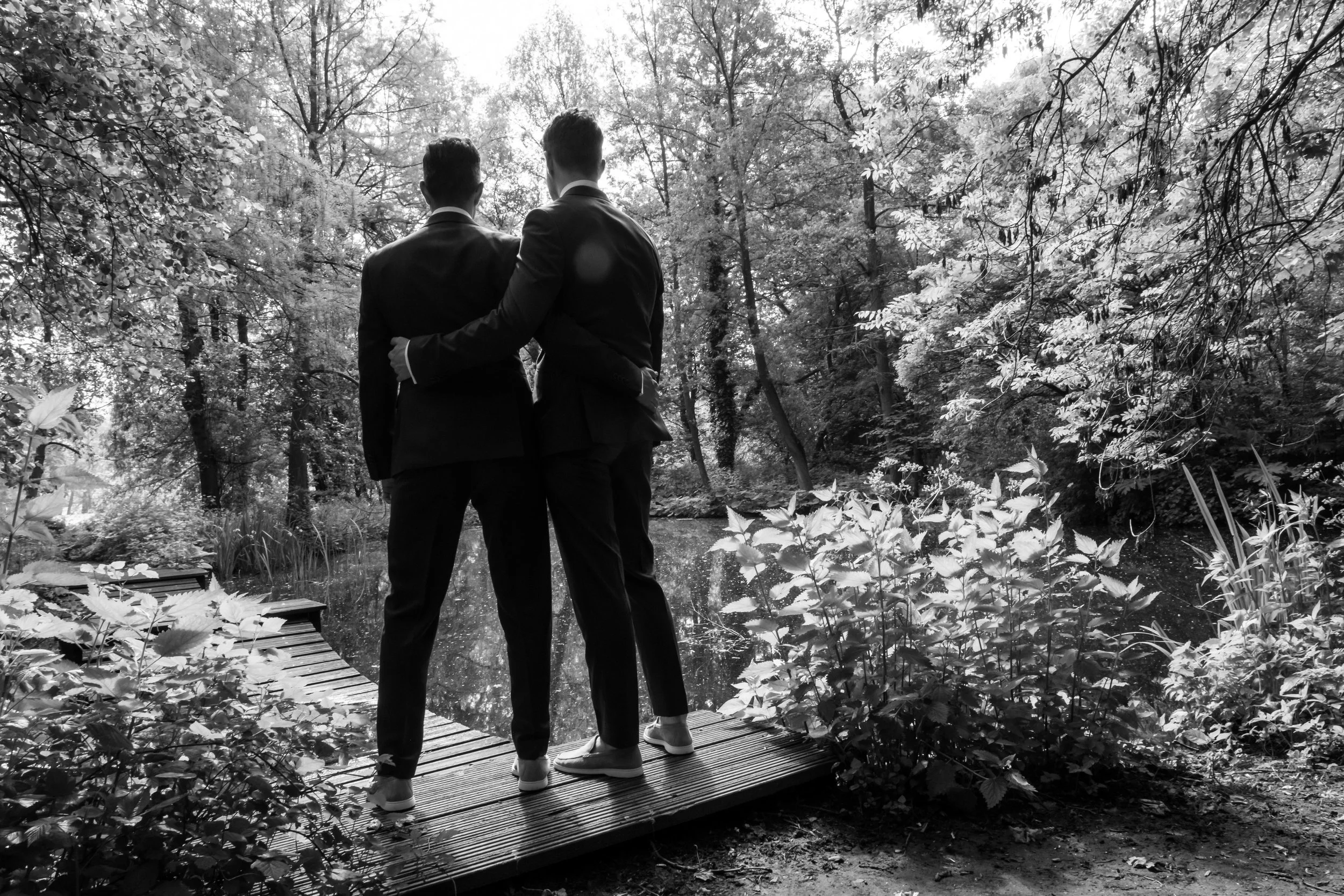 Twee mensen in pakken die elkaar vasthouden op een houten pad in een bos, zwart-wit foto.