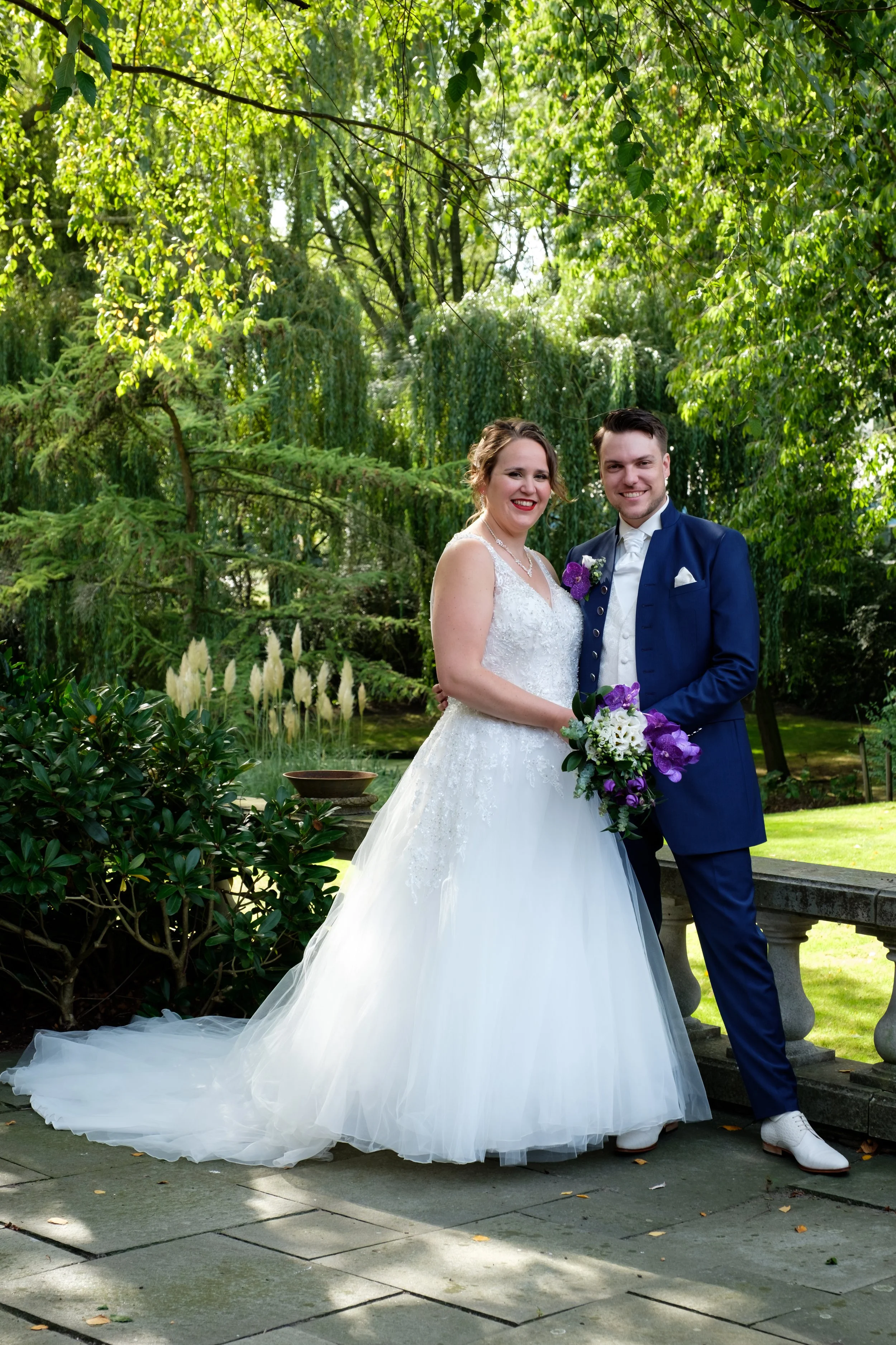 Een bruid en bruidegom poseren samen in een tuin met groen bladerendek. De bruid draagt een witte jurk met tule en kant, en houdt een boeket met witte en paarse bloemen. De bruidegom draagt een donkerblauw pak en witte schoenen.