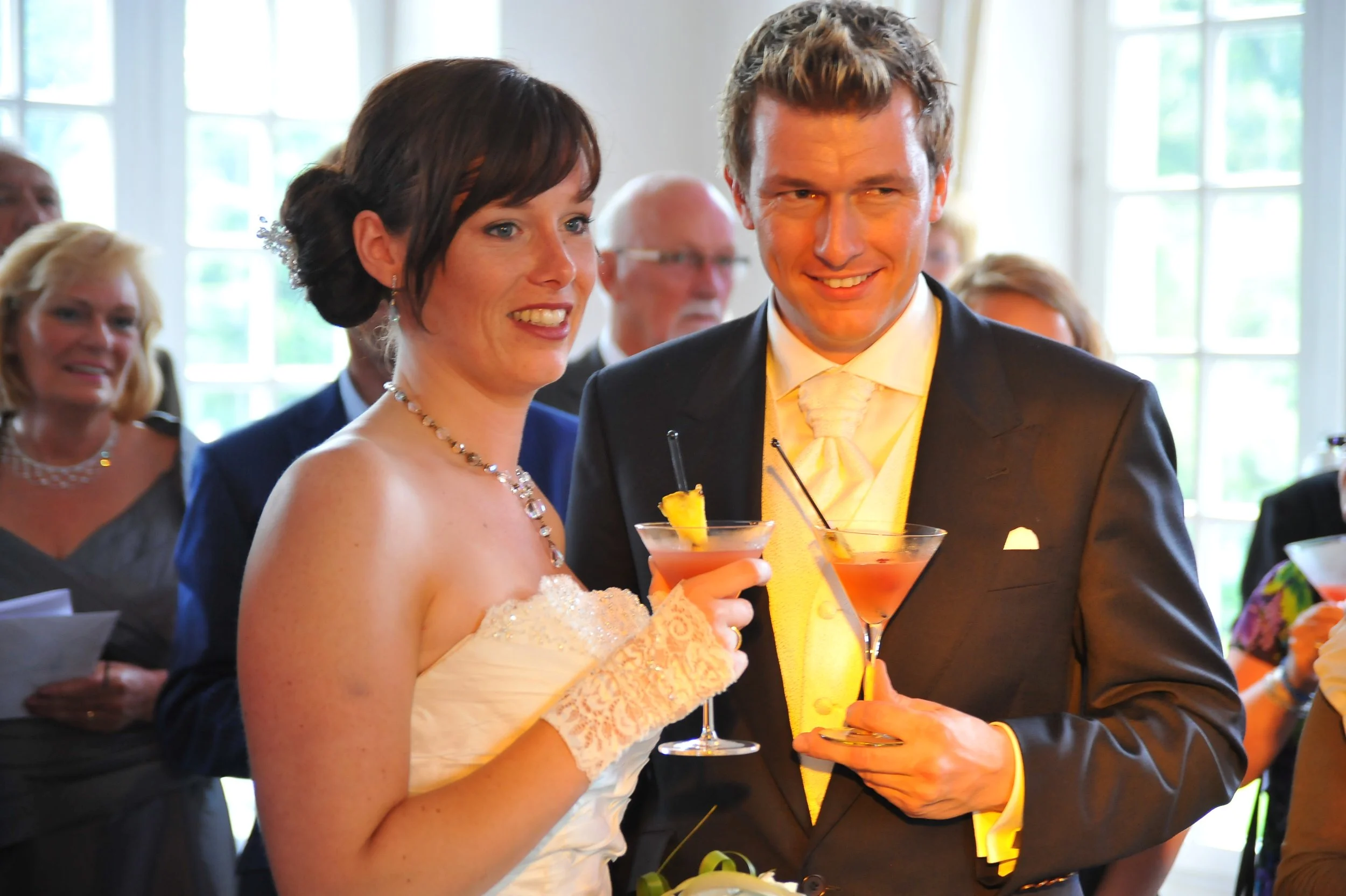 Bruid en bruidegom tijdens een feest, beiden lachen en houden drankjes vast. De sfeer is feestelijk en de gasten zijn zichtbaar op de achtergrond.