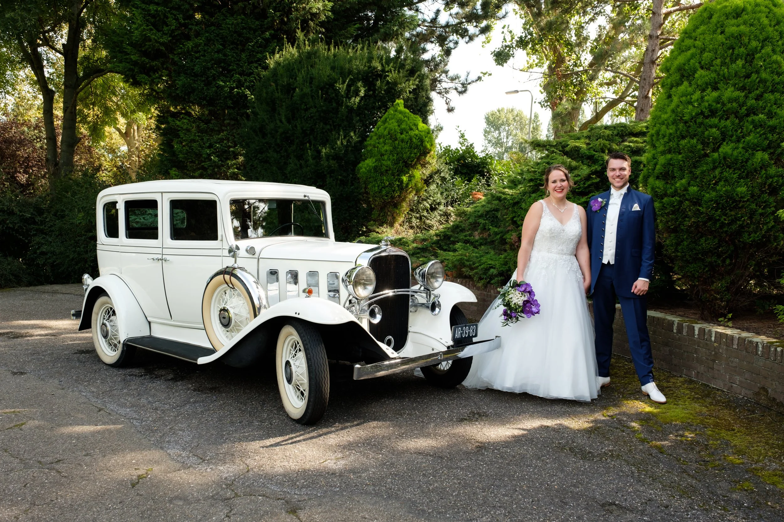 Een bruid en bruidegom in een tuxedo en een trouwjurk staat naast een witte vintage auto in een groene tuin.