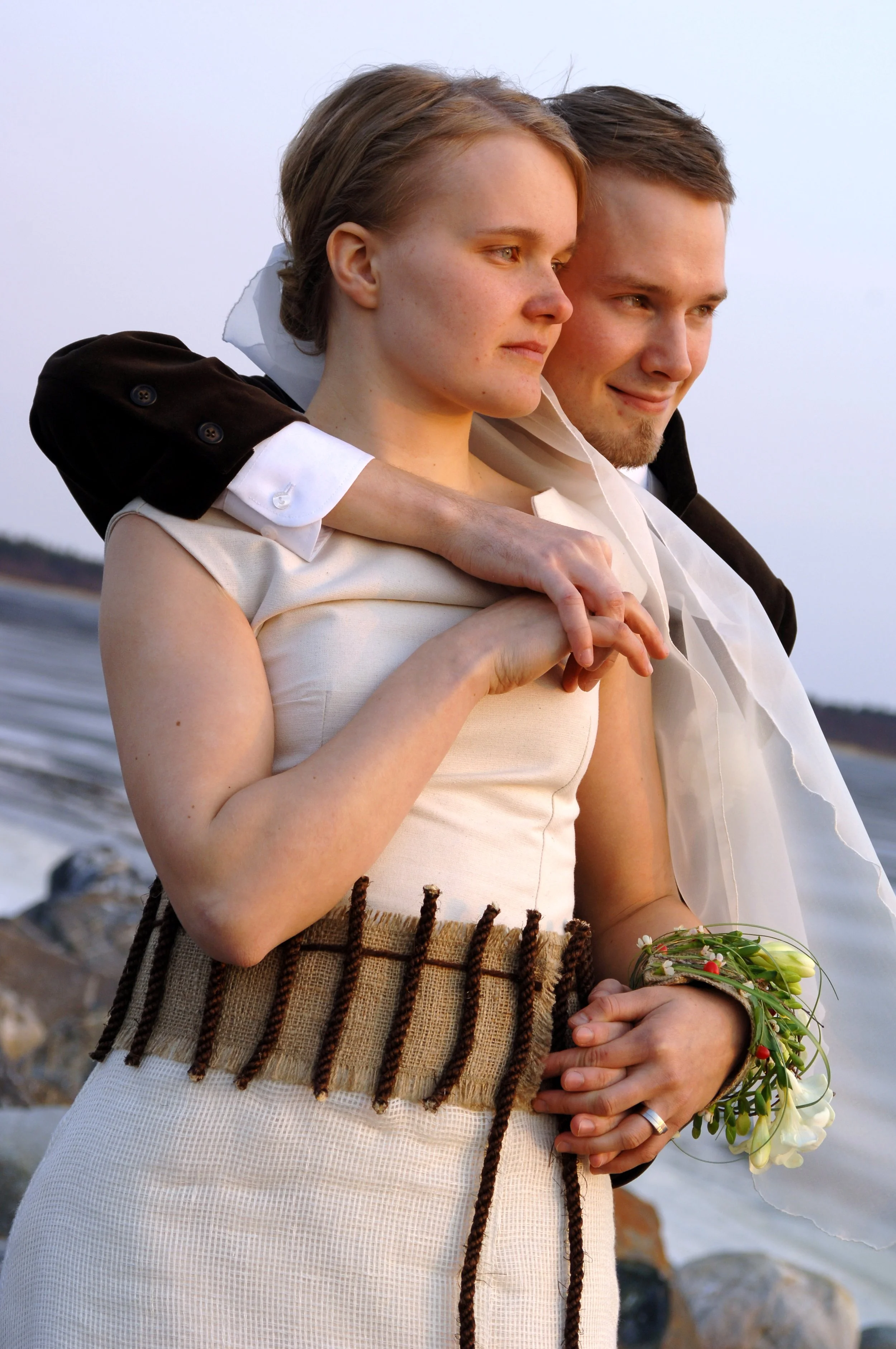 Een close-up van een vrouw en een man die dicht bij elkaar staan, met de zee en rotsen op de achtergrond. Ze lijken gelukkig en intiem, met de vrouw die een boeket bloemen vasthoudt.