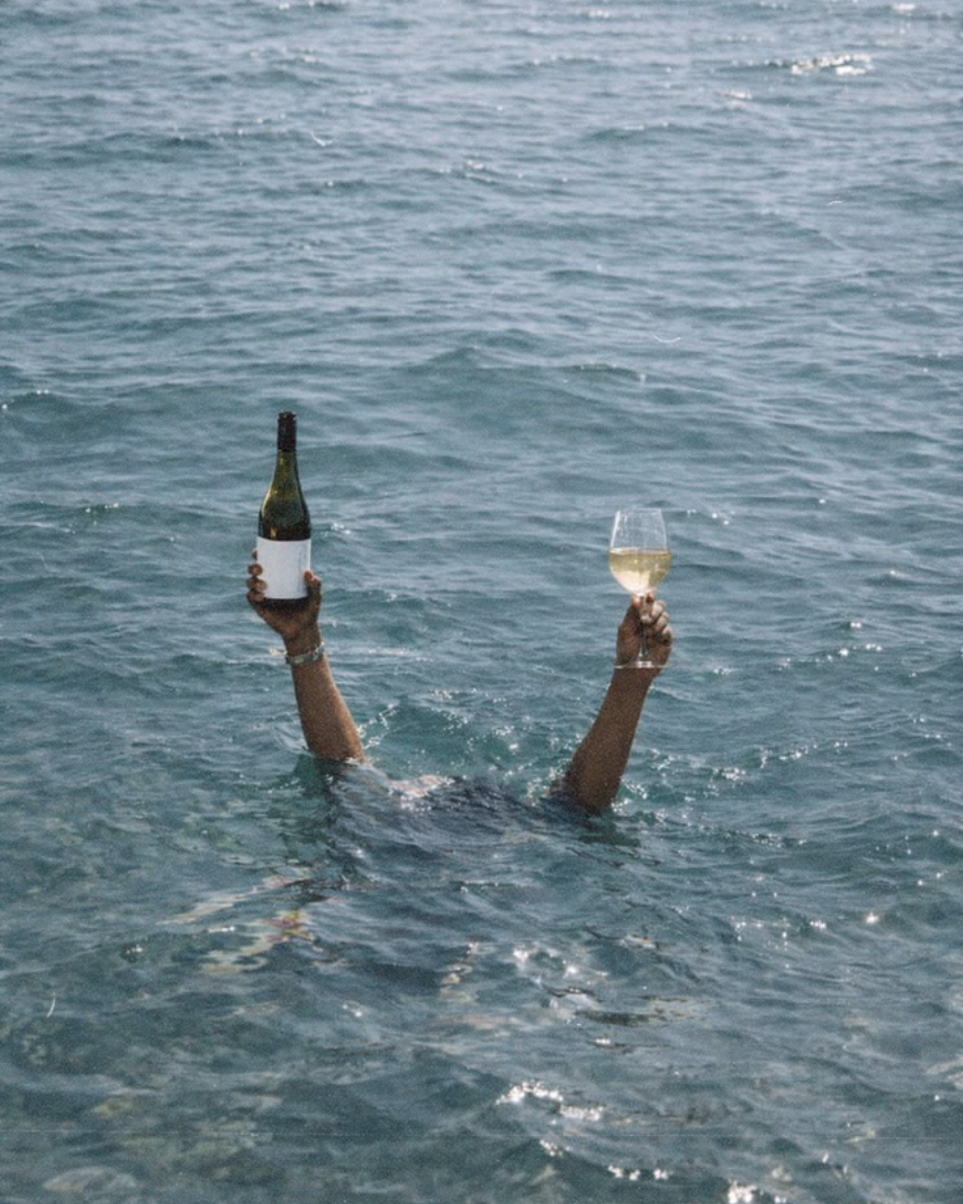 Two hands emerging from the water holding a bottle of wine and a glass of white wine, with the ocean in the background